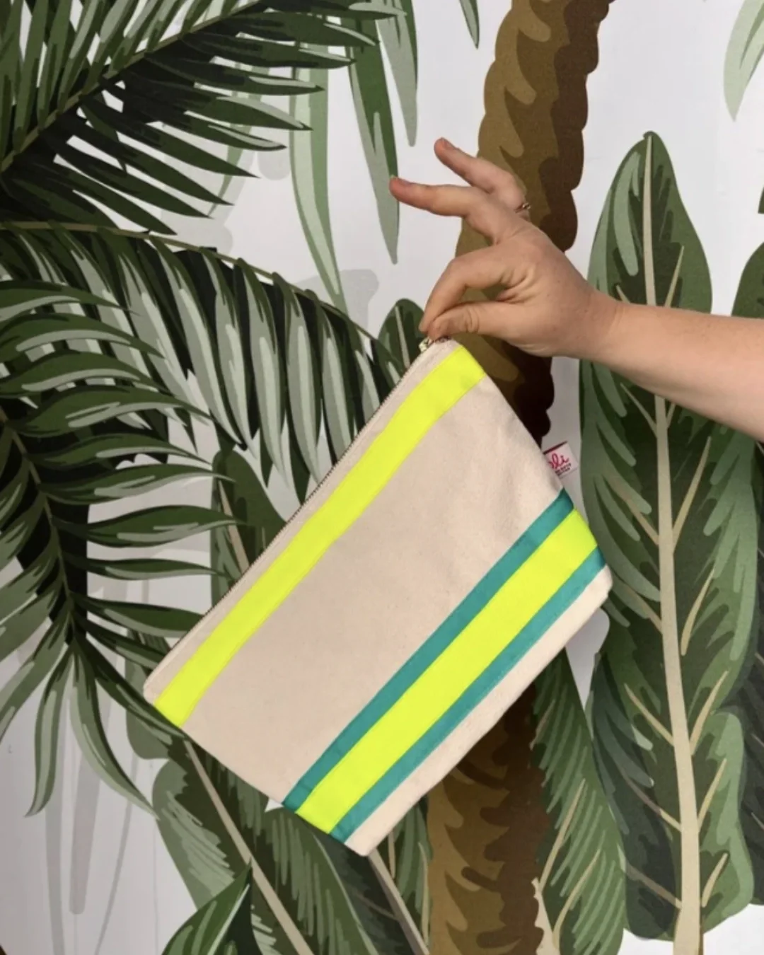 A hand holding a cream cotton pouch with bright neon yellow and teal green stripes against a tropical palm leaf backdrop, handmade by Soli in Cape Town for retail sustainable merchandise.