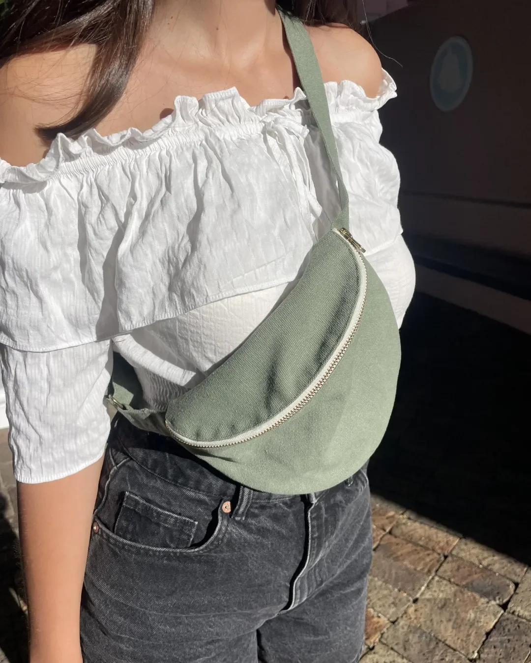 A person wearing an olive green handmade cotton canvas moon bag across a white ruffle top and black jeans, highlighting a sustainable fanny pack design by Soli in Cape Town for corporate gifts and merchandise. 