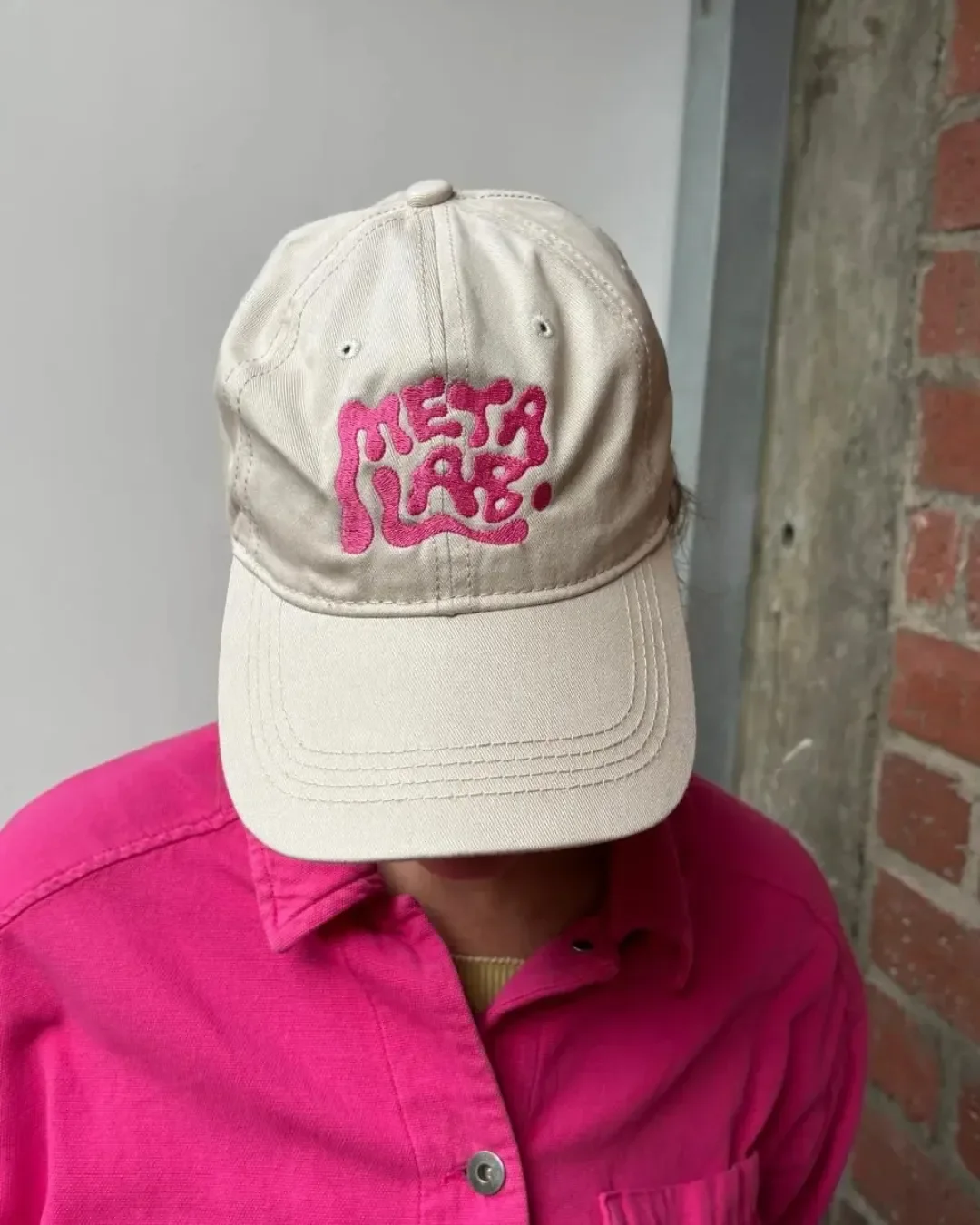 Rear view of a custom cream-colored dad cap featuring a small pink and green cartoon character embroidered on the back, worn by a person with their hair styled in a bun with a leopard print scrunchie, paired with a bright pink jacket.