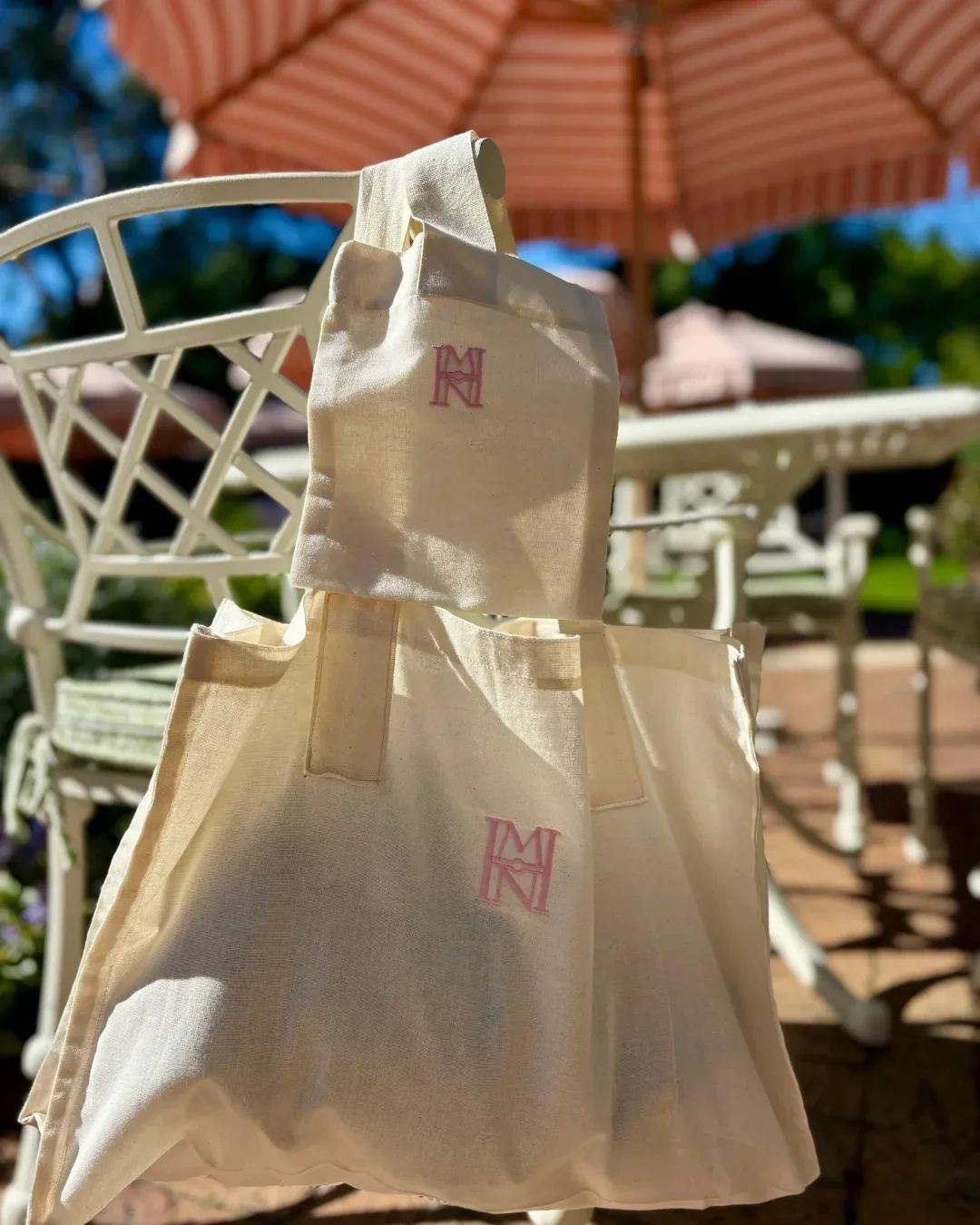 The Mount Nelson Gift Bag set in light natural calico featuring the signature pink embroidered MN logo hanging in a sunlit outdoor garden setting, Handmade by Soli in Cape Town for sustainable merchandise or corporate gifts.