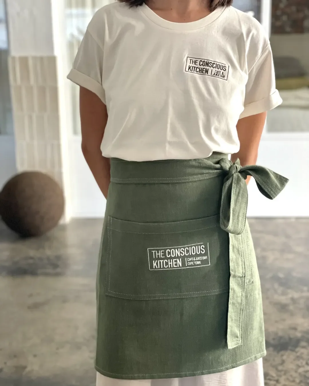 A woman wearing an olive green sustainable canvas half apron with "The Conscious Kitchen" logo, handmade by local artisans in Cape Town for hospitality use.
