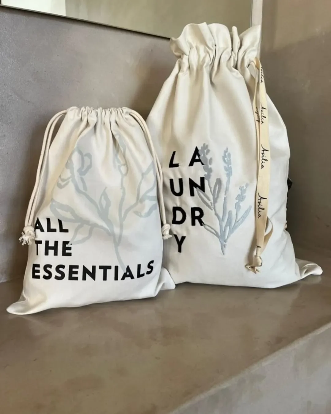 Set of two custom branded off-white cotton hospitality drawstring bags featuring LAUNDRY and ALL THE ESSENTIALS text with botanical prints by Soli Cape Town