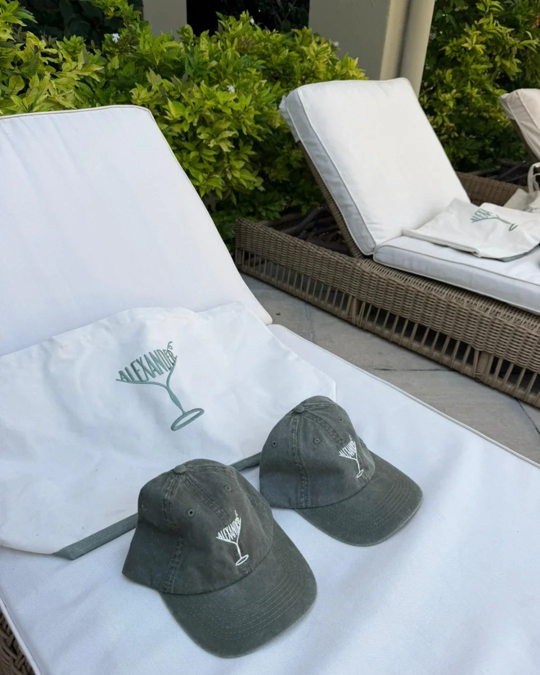 Two olive green caps and a cream cotton canvas Pezula bag with custom "ALEXANDER" martini glass embroidery resting on white outdoor lounge chairs, handmade by Soli in Cape Town for sustainable merchandise or corporate gifts.