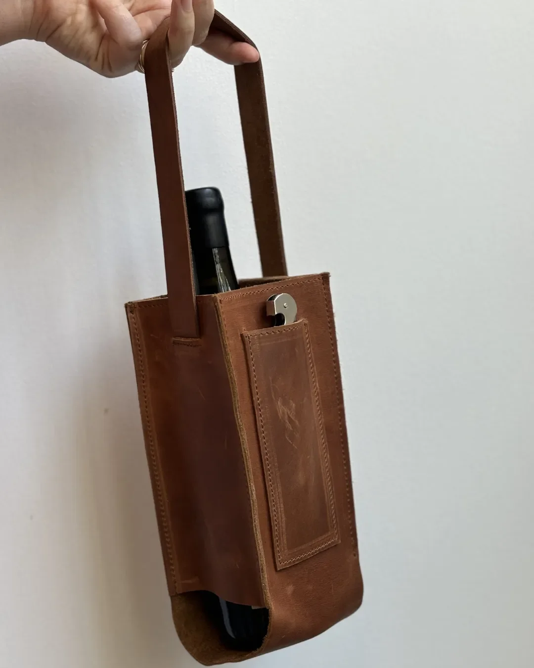A person holding a premium handmade brown leather wine carrier with a single handle, carrying a wine bottle and a stainless steel corkscrew, Handmade by Soli in Cape Town for sustainable merchandise or corporate gifts.