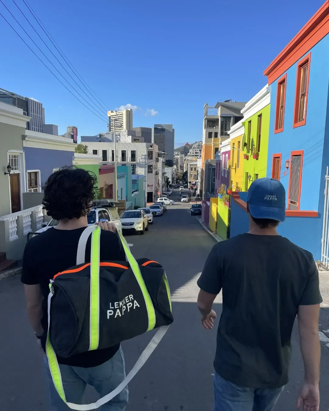 Two men walking through the colorful streets of Bo-Kaap with a black canvas duffel bag and a blue cap, handmade by Soli in Cape Town for sustainable retail merchandise.