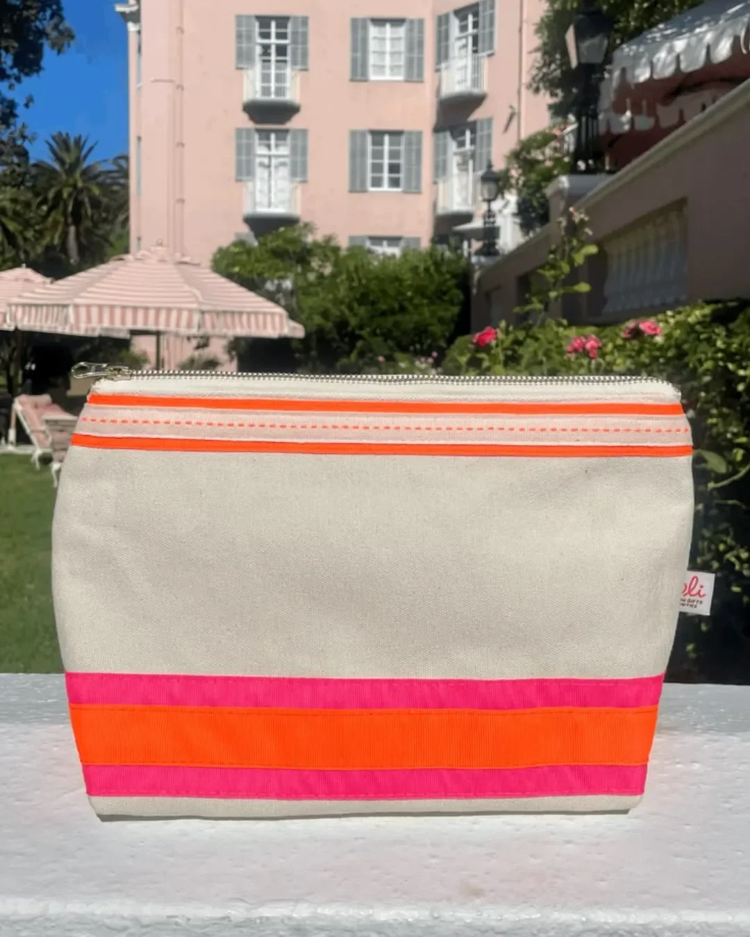 A woman in a sun hat and sunglasses holding a cream cotton pouch with neon pink and orange stripes in front of a colorful mural, handmade by Soli in Cape Town for sustainable merchandise.