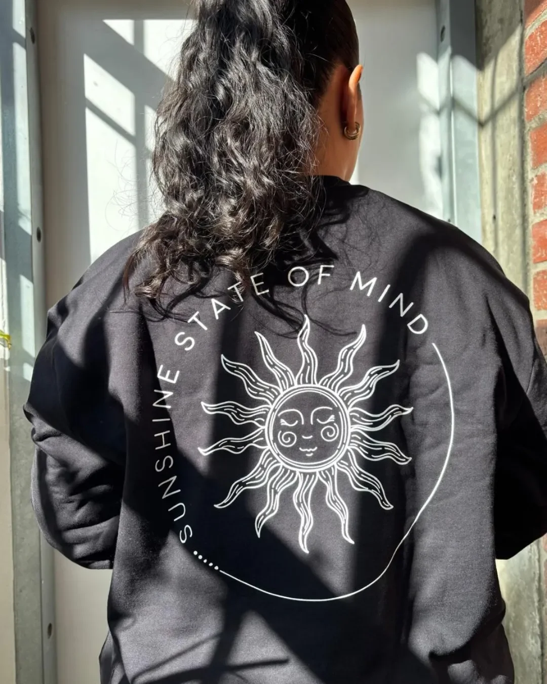 Back view of a custom black sweatshirt featuring a white printed graphic of a smiling sun and the phrase "SUNSHINE STATE OF MIND", custom merchandise made by Soli.