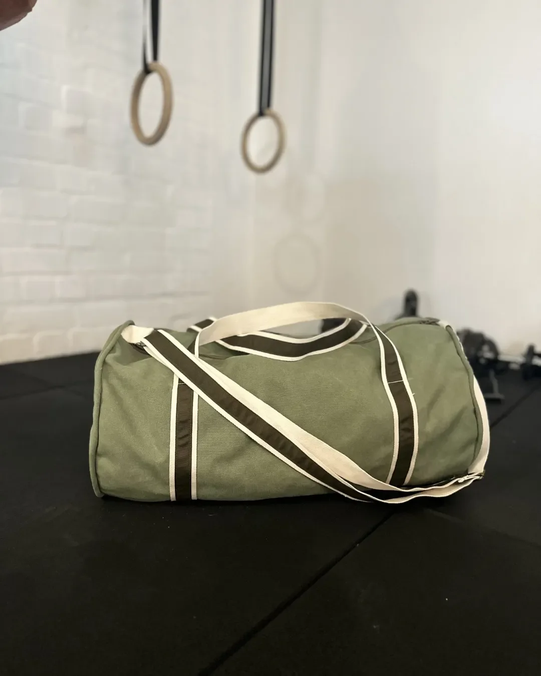 An olive green canvas gym duffel bag with white and brown striped straps resting on a black gym mat with gymnastic rings in the background, handmade by Soli in Cape Town for sustainable retail merchandise.
