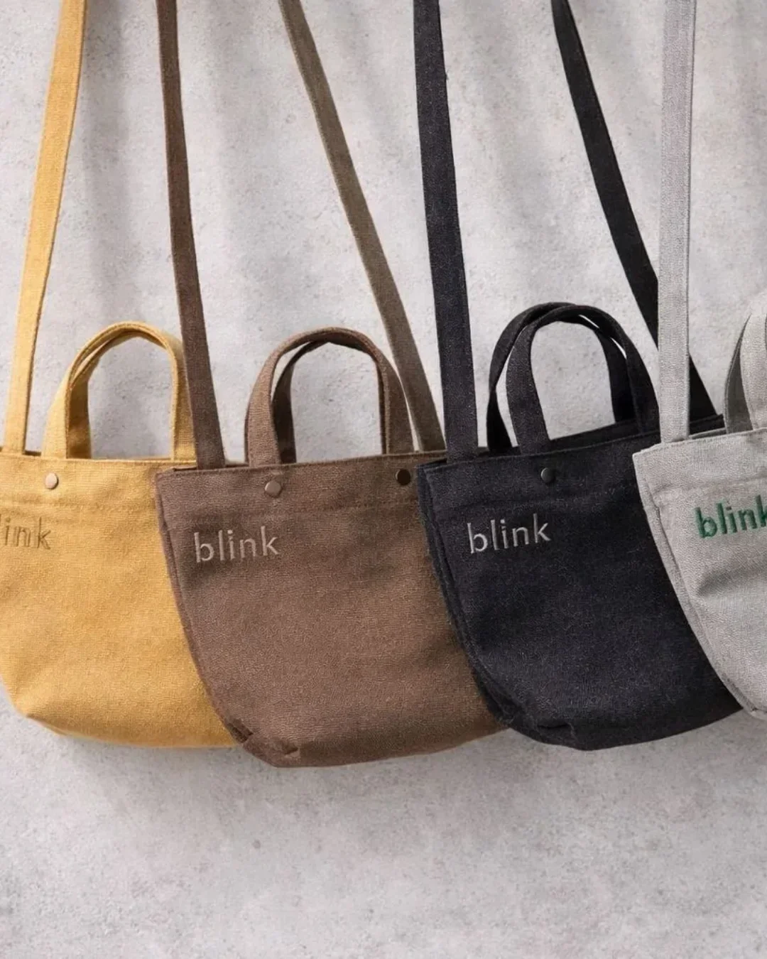 A row of four Japanese-style wrap bags in yellow, brown, charcoal, and grey featuring custom "blink" embroidery and long shoulder straps against a concrete wall, handmade by Soli in Cape Town for sustainable merchandise or corporate gifts.