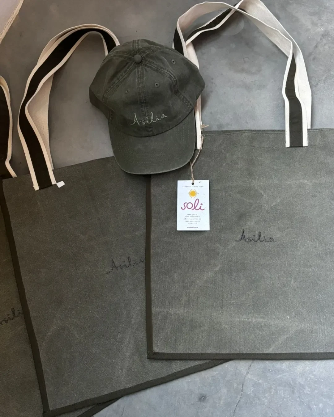 Flat lay of handmade olive green tote bags with custom embroidery alongside a branded Soli tag, showcasing custom sustainable corporate gifts from Cape Town.