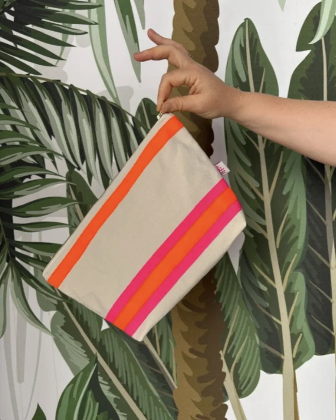 A hand holding a cream cotton pouch with vibrant neon pink and orange stripes against a tropical palm leaf background, handmade by Soli in Cape Town for sustainable retail merchandise.