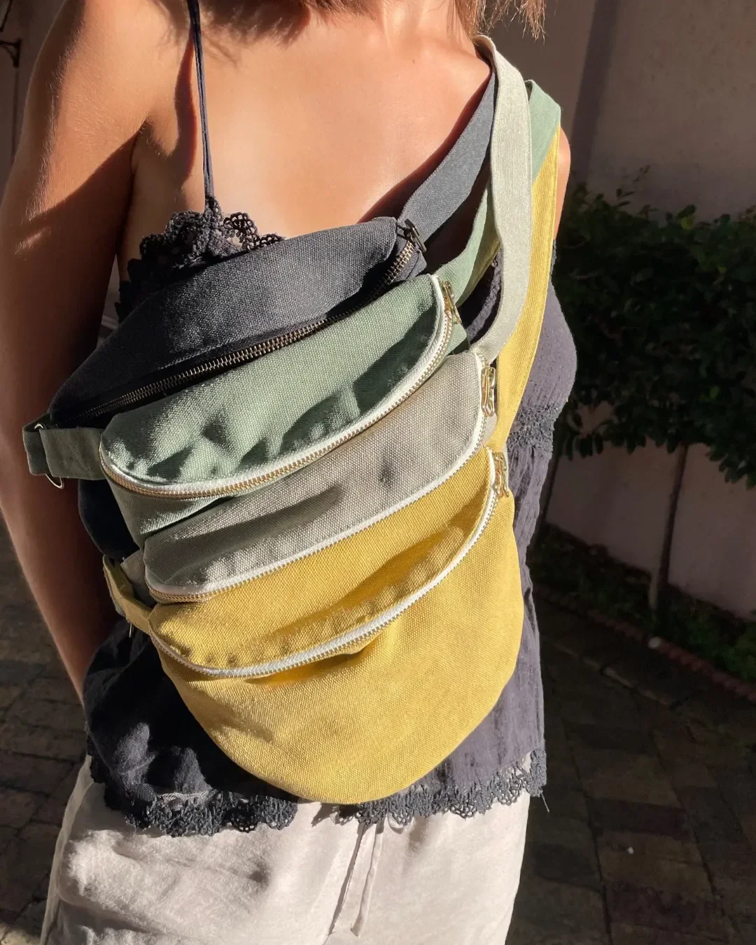 A person wearing a stack of four handmade cotton canvas moon bags in charcoal, sage green, light grey, and mustard yellow, showcasing the variety of colors available for custom sustainable merchandise in cape town.