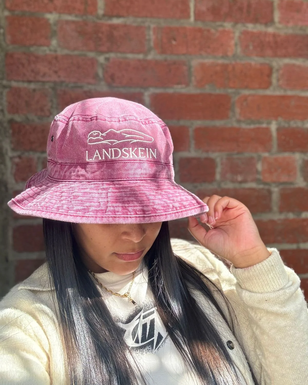 A person wearing a pink washed canvas bucket hat embroidered with the white Landskein logo and silhouette, posing against a brick wall background for custom corporate gifts.