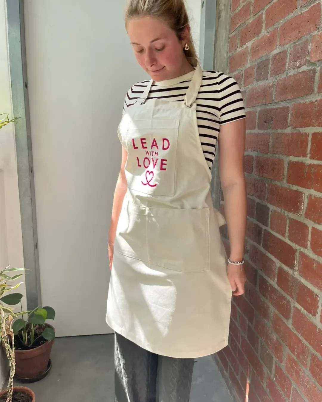 Custom branded cream canvas full body apron featuring pink 'Lead With Love' embroidery, handmade by Soli's local artisans in Cape Town for hospitality apron merchandise for staff.