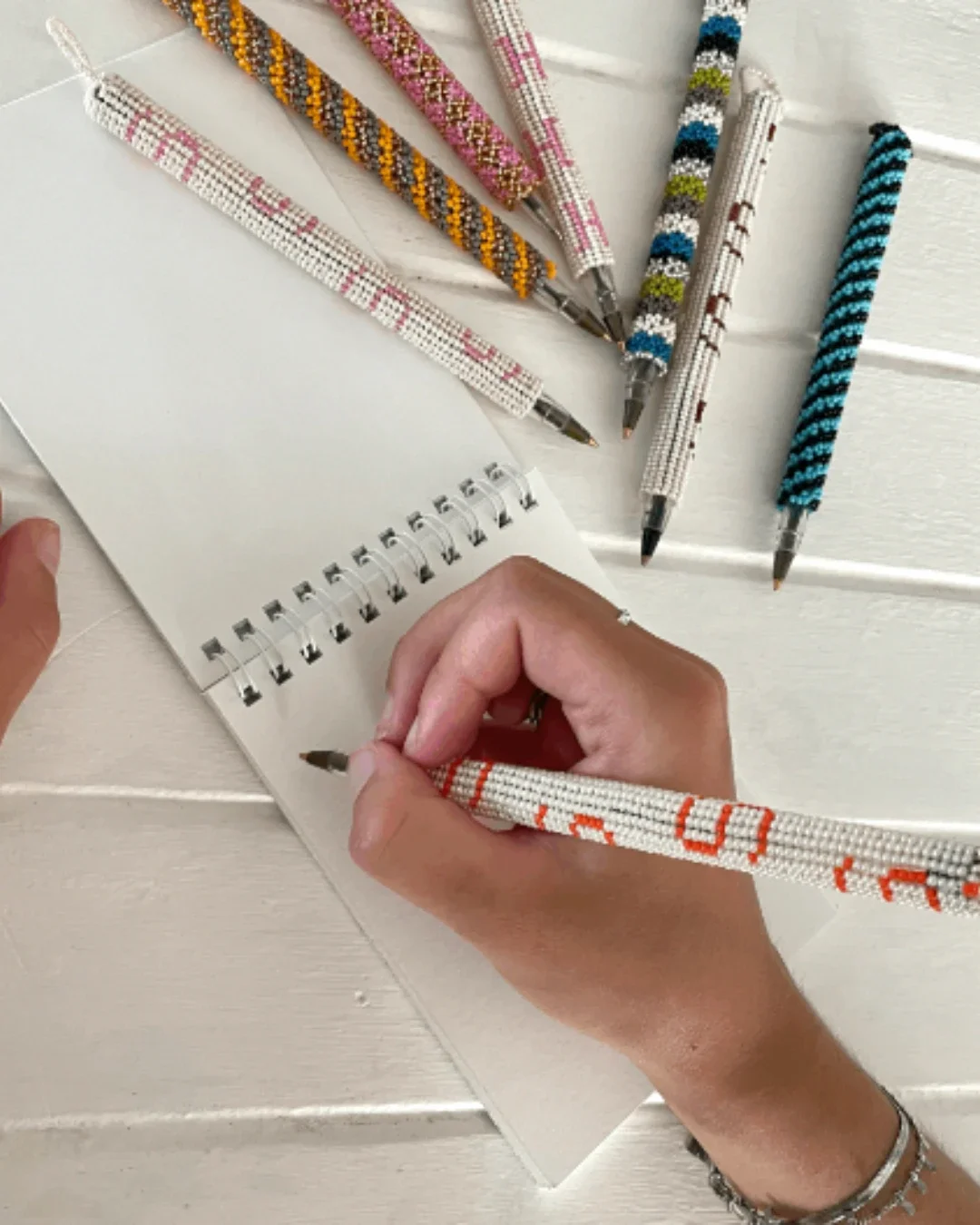 A variety of colorful beaded pens with intricate geometric patterns and initials being used to write in a notebook, Handmade by Soli in Cape Town for sustainable merchandise or corporate gifts.