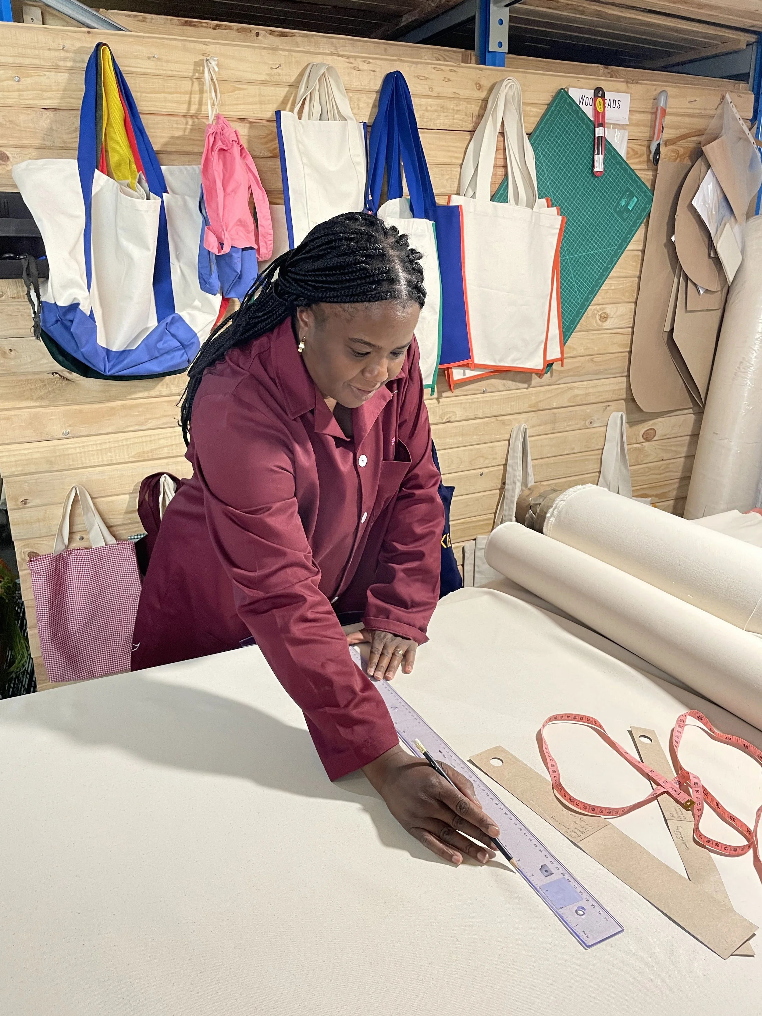 Person measuring fabric with a ruler on a sewing table, surrounded by tote bags hanging on a wooden wall.