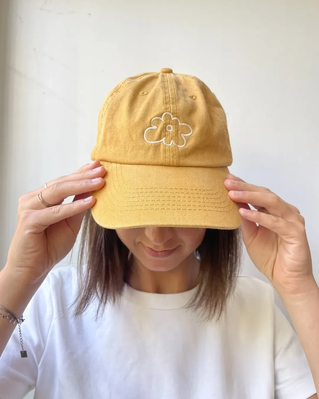 A person wearing a mustard yellow vintage-style washed cotton cap with a white embroidered flower logo, part of the Soli custom branded headwear collection. Perfect for sustainable branded merchandise, locally finished by South African artisans.