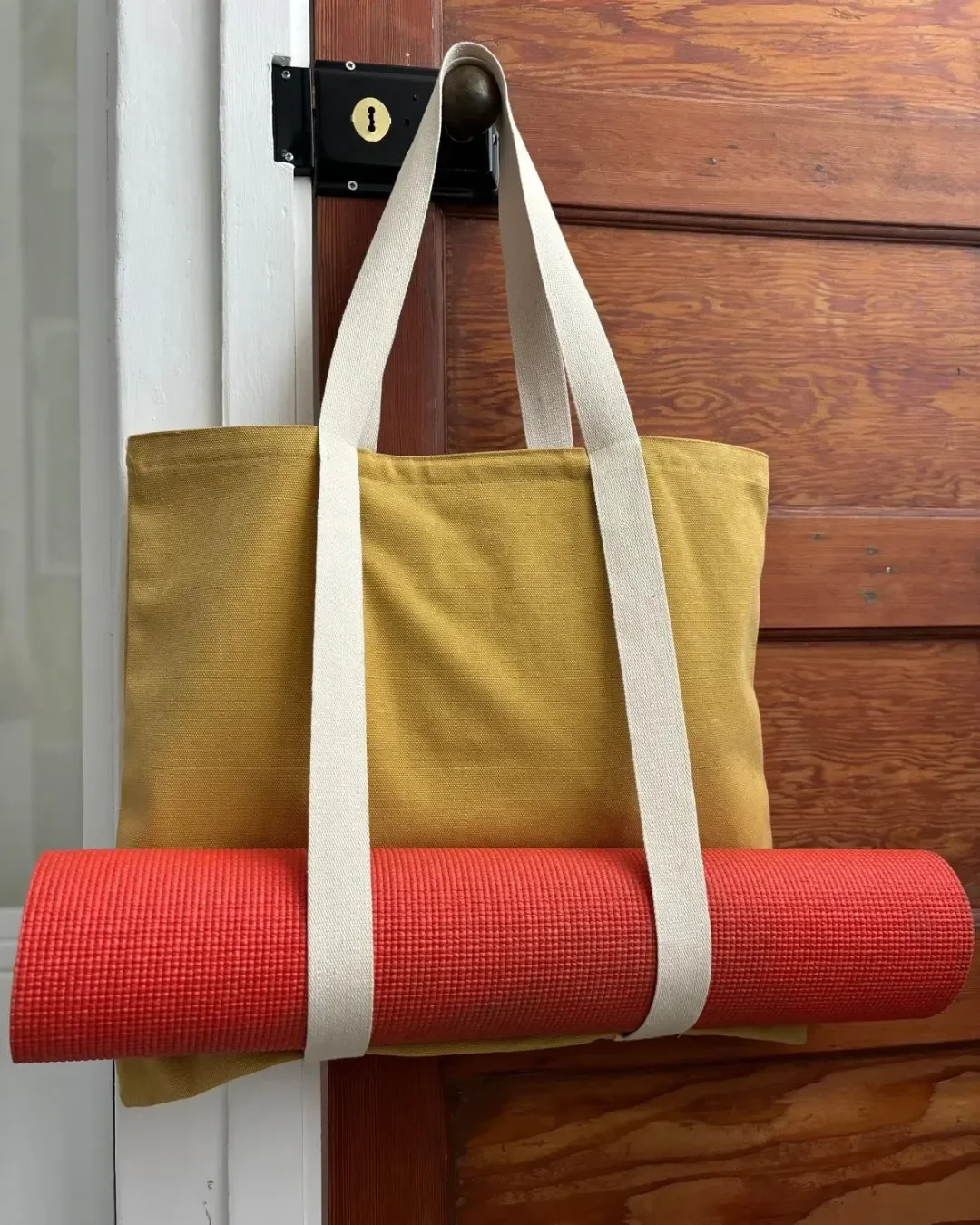 Mustard yellow heavyweight canvas Vienna Tote hanging on a wooden door, featuring integrated cotton straps holding a red yoga mat, handmade in Cape Town by Soli for a custom yoga or pilates bag merchandise or corporate gift.