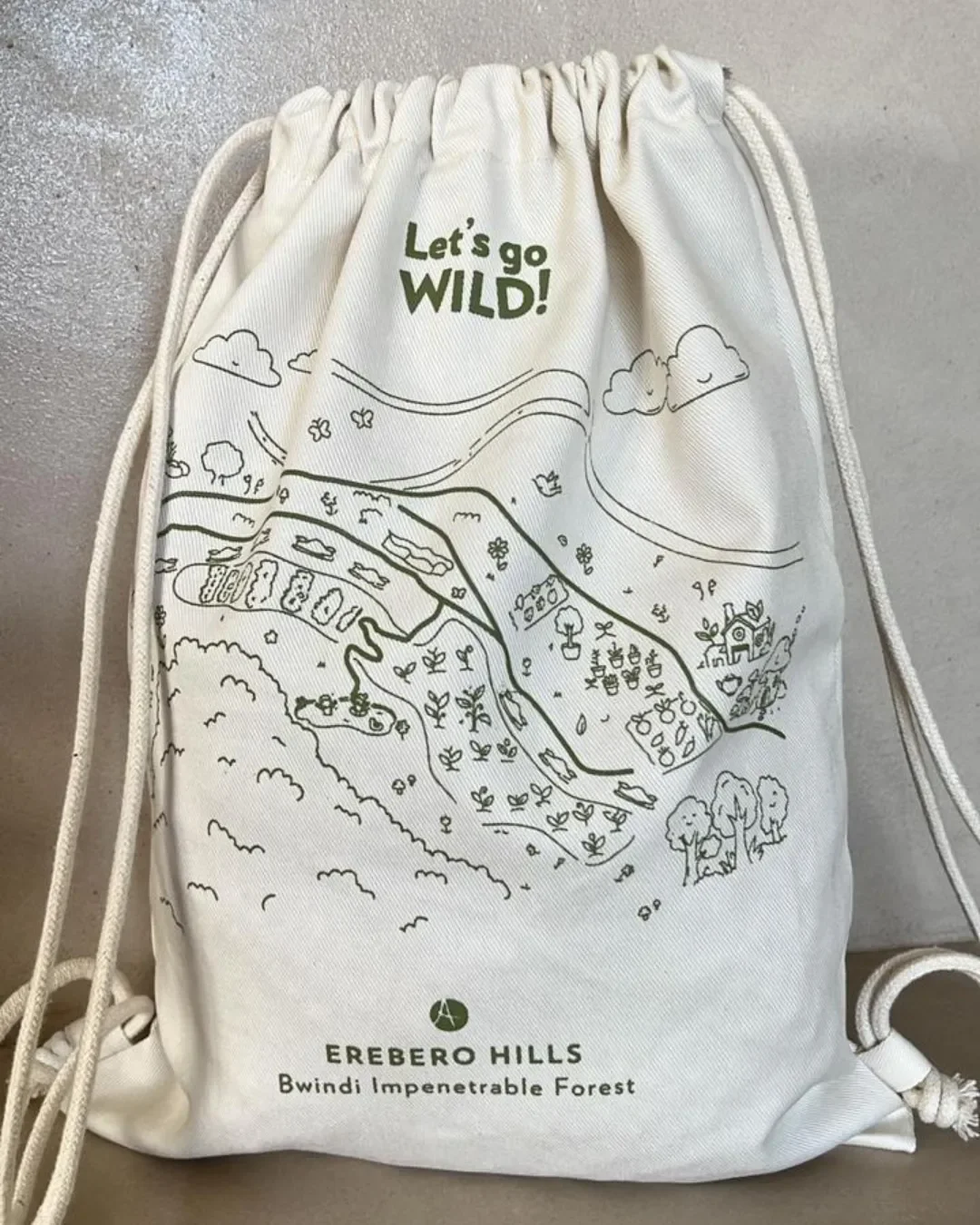 Natural cream cotton drawstring backpack featuring a green illustrated landscape map with "Let's go WILD!" and "EREBERO HILLS Bwindi Impenetrable Forest" printed at the bottom, custom made by Soli.