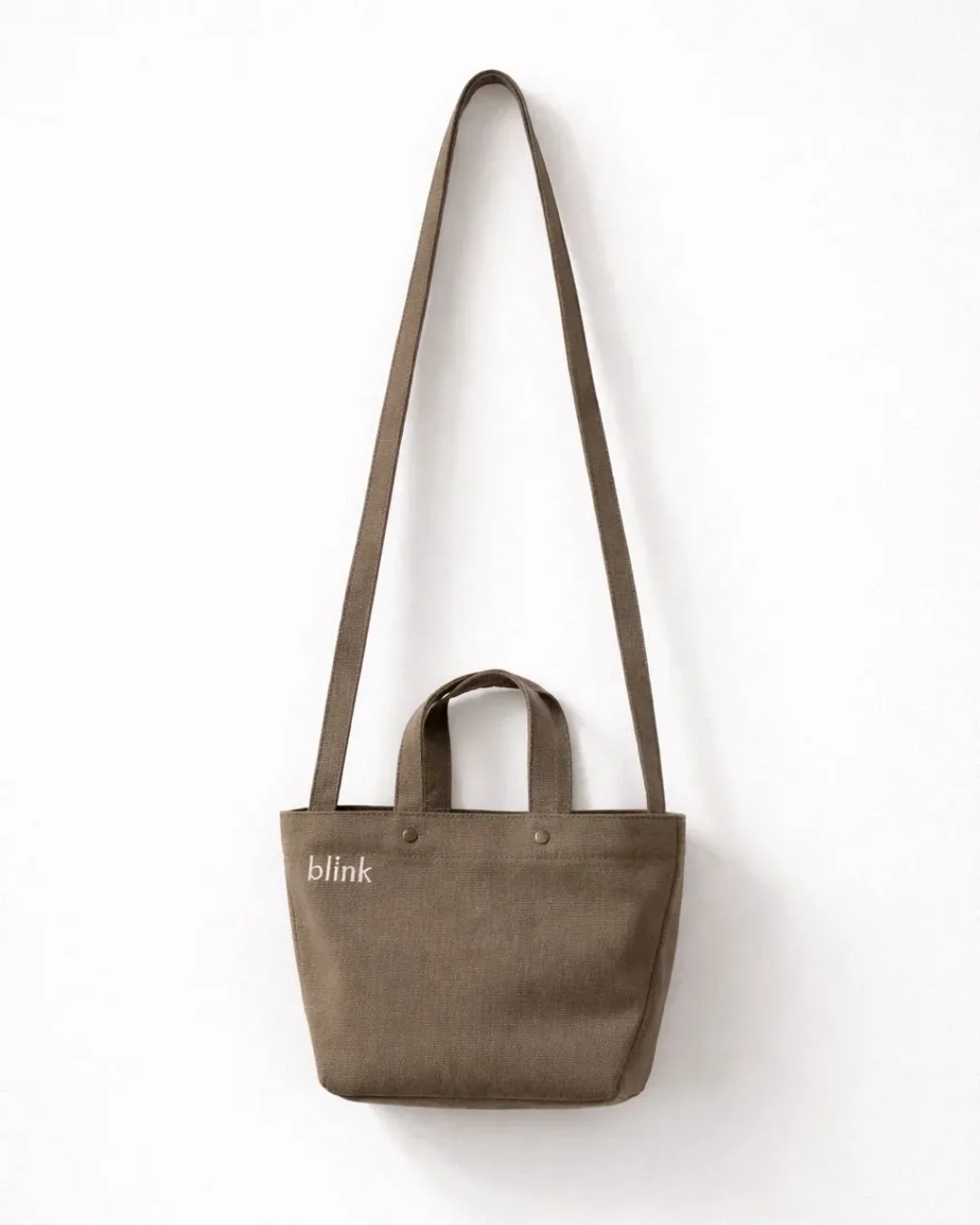 A durable brown heavy-duty fabric Japanese-style wrap bag featuring a long shoulder strap and custom "blink" embroidery hanging against a white wall, handmade by Soli in Cape Town for sustainable merchandise or corporate gifts.