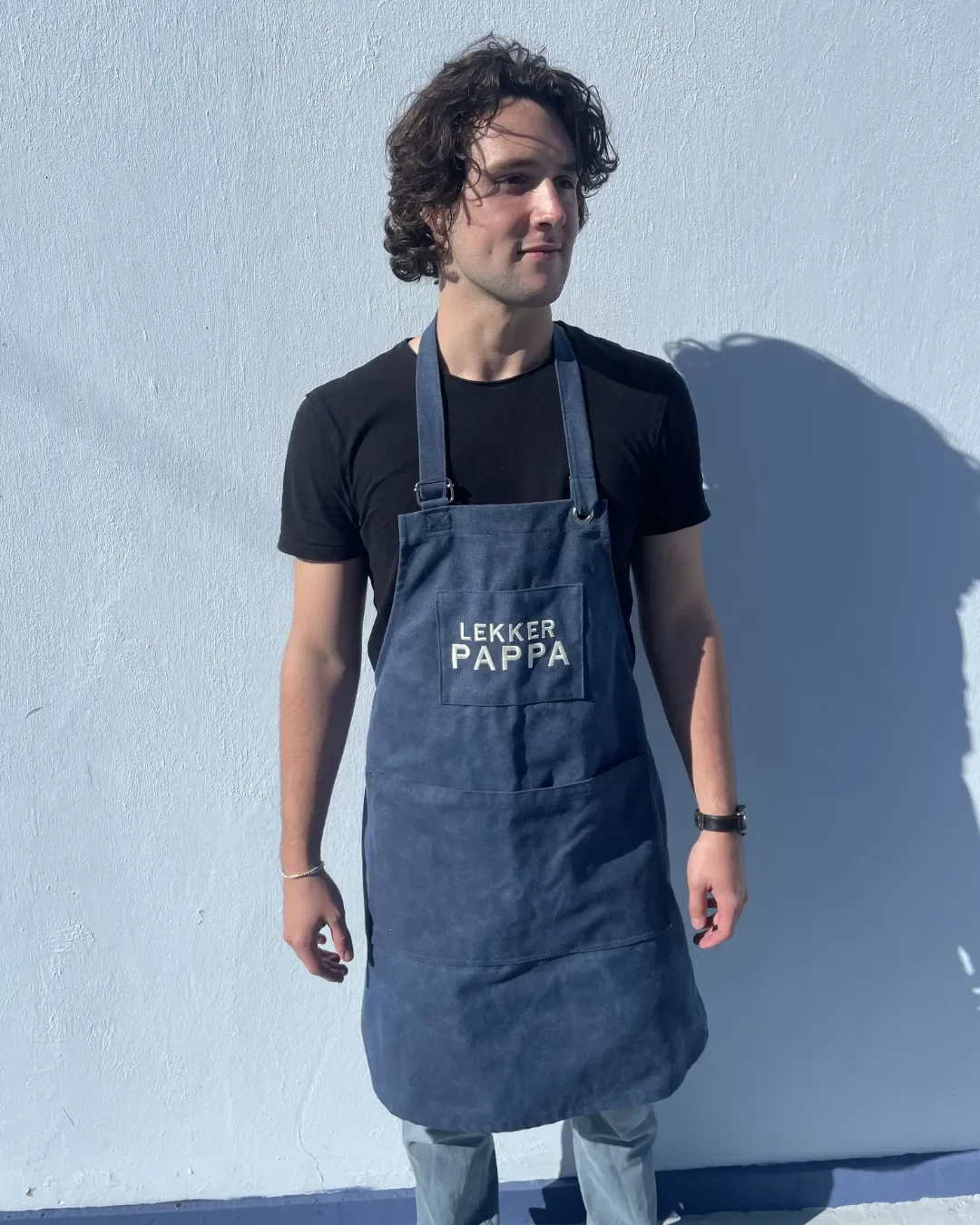A male model wearing a full-length blue canvas hospitality apron featuring a custom embroidered logo, handmade by local artisans in Cape Town by soli.