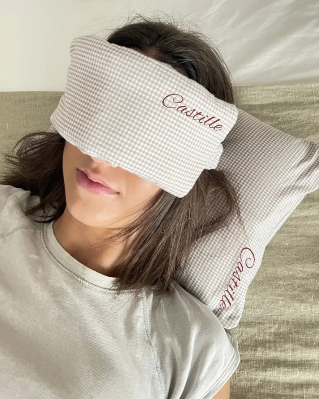 A person using a matching gingham check travel eye mask and pillow set with custom "Castille" embroidery, handmade from 100% local cotton by Soli in Cape Town.