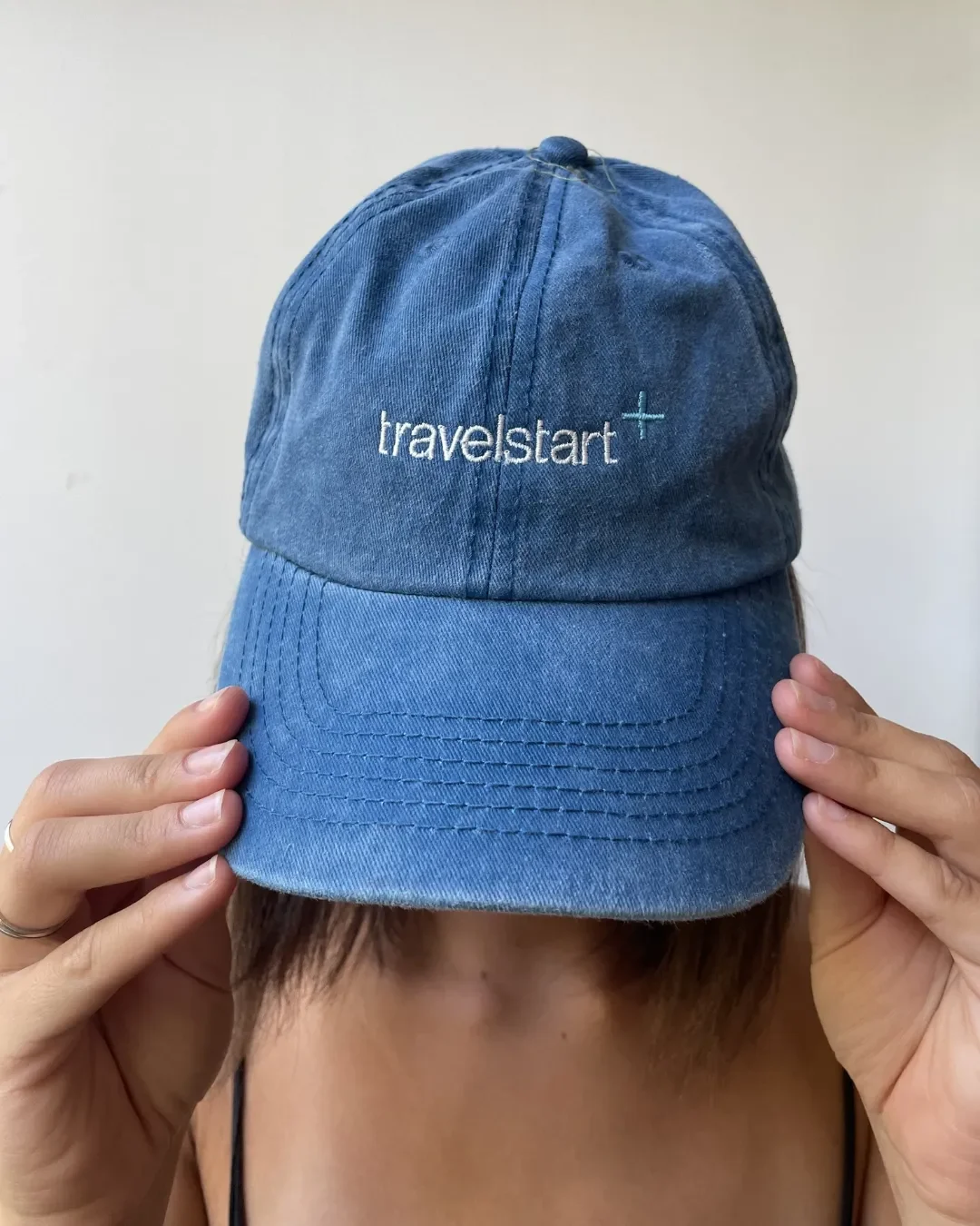 Front view of a washed denim blue vintage style cotton cap featuring custom white and turquoise embroidery for Travelstart, held by a person. Fully customizable with printed or embroidered logos, these one-size-fits-all caps are perfect for premium c