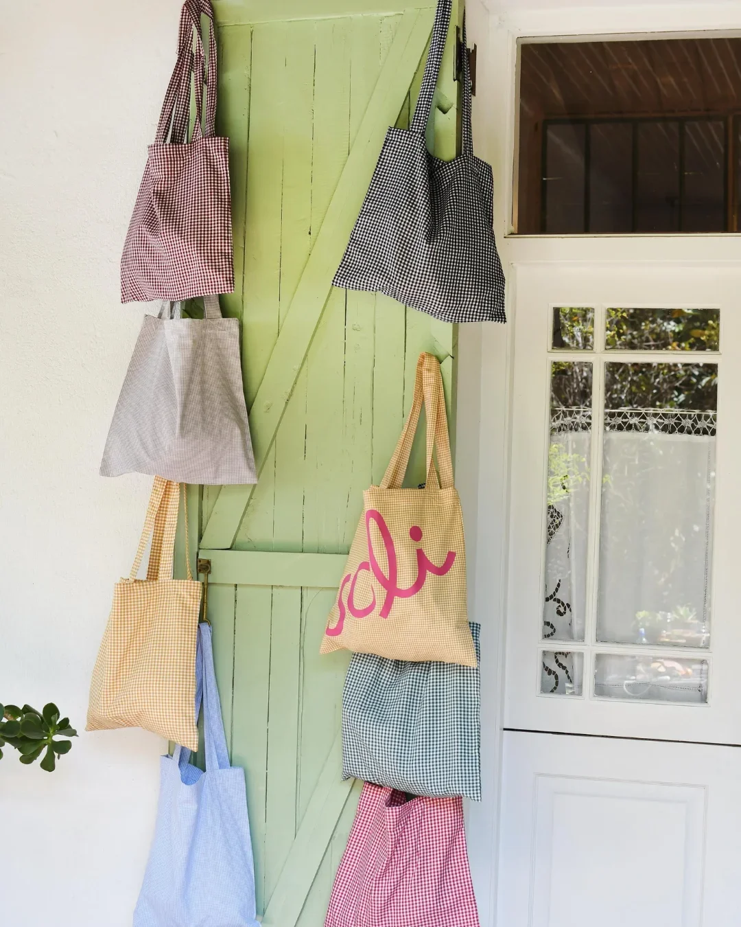 A collection of Frenchy Totes in various checkered gingham colors including yellow, pink, blue, and maroon hanging on a light green wooden door, handmade by Soli artisans in Cape Town.