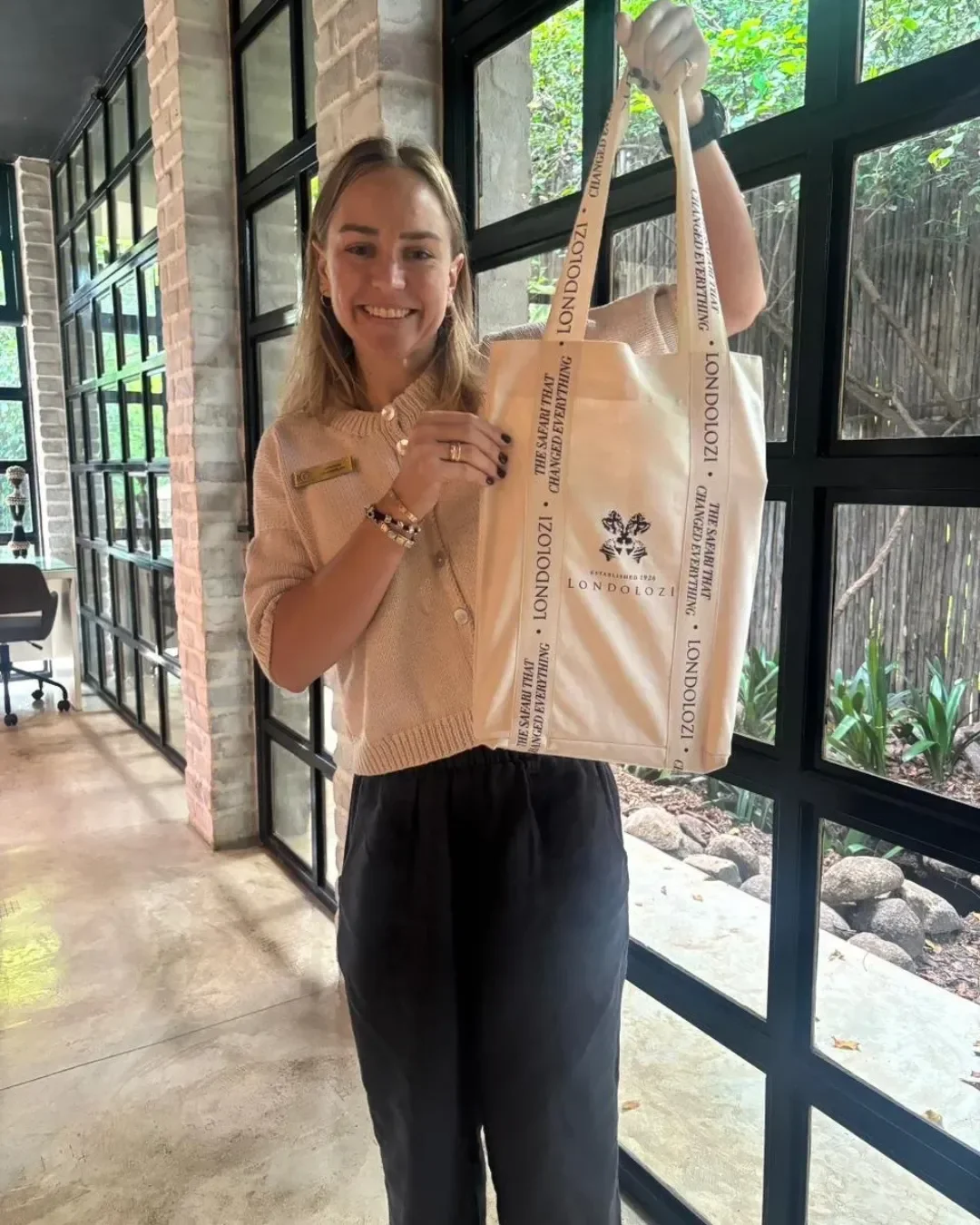 A smiling woman holding up a premium cream canvas signature strap tote bag branded with the Londolozi logo and custom printed text on the handles, showcasing luxury safari lodge corporate merchandise.