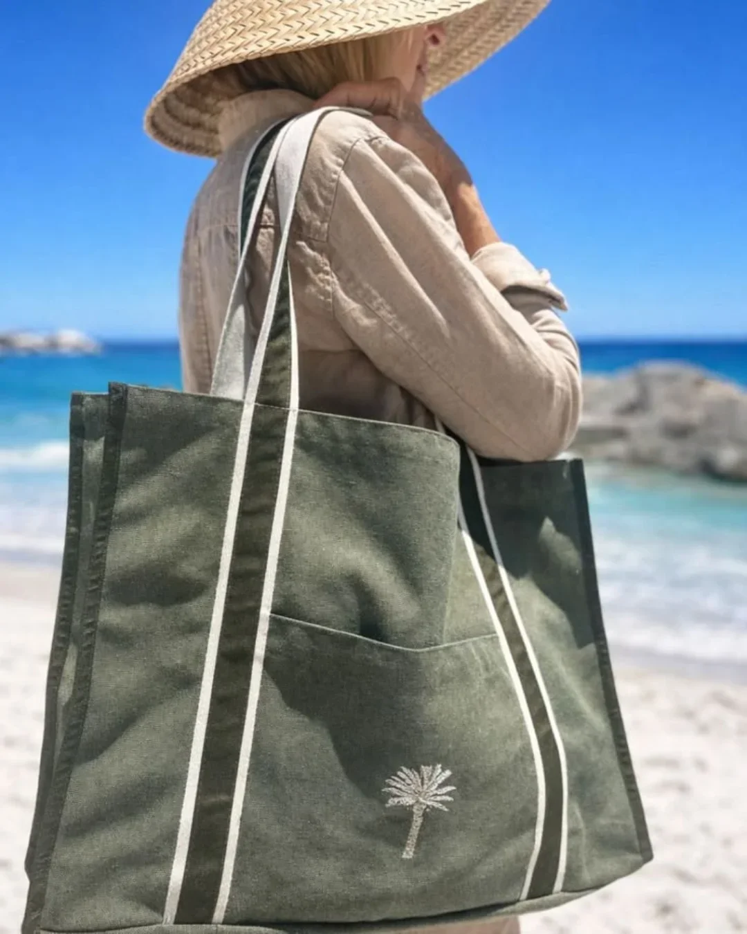 A woman at the beach carrying an olive green heavy canvas Diesel Bag with cream contrast handles and a white embroidered palm tree logo, Handmade by Soli in Cape Town for merchandise or corporate gifts.