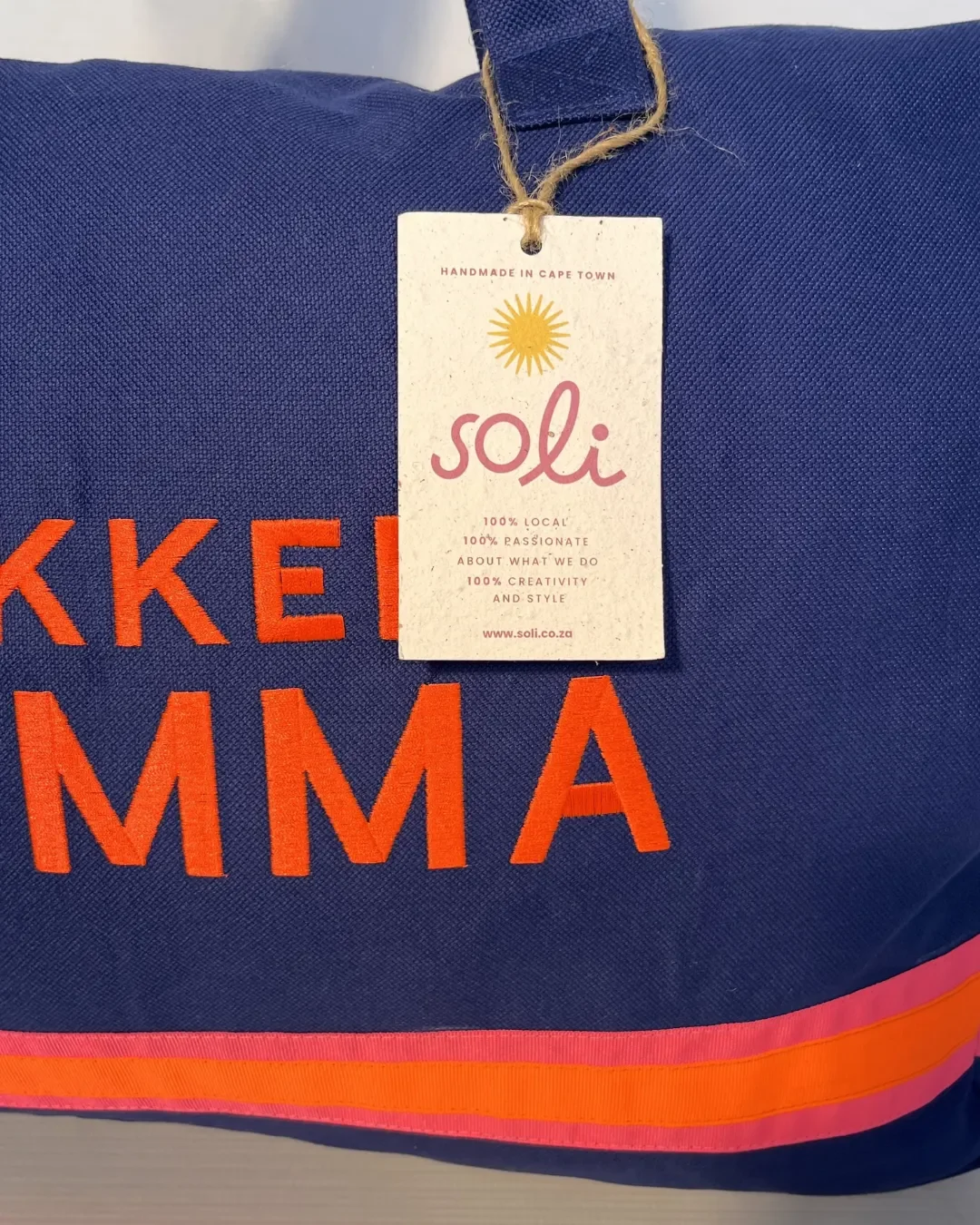 A close-up of a navy blue Lekker Mamma tote bag featuring a "Soli" brand tag tied with twine, showing high-quality orange embroidery, handmade by Soli in Cape Town for sustainable retail merchandise.