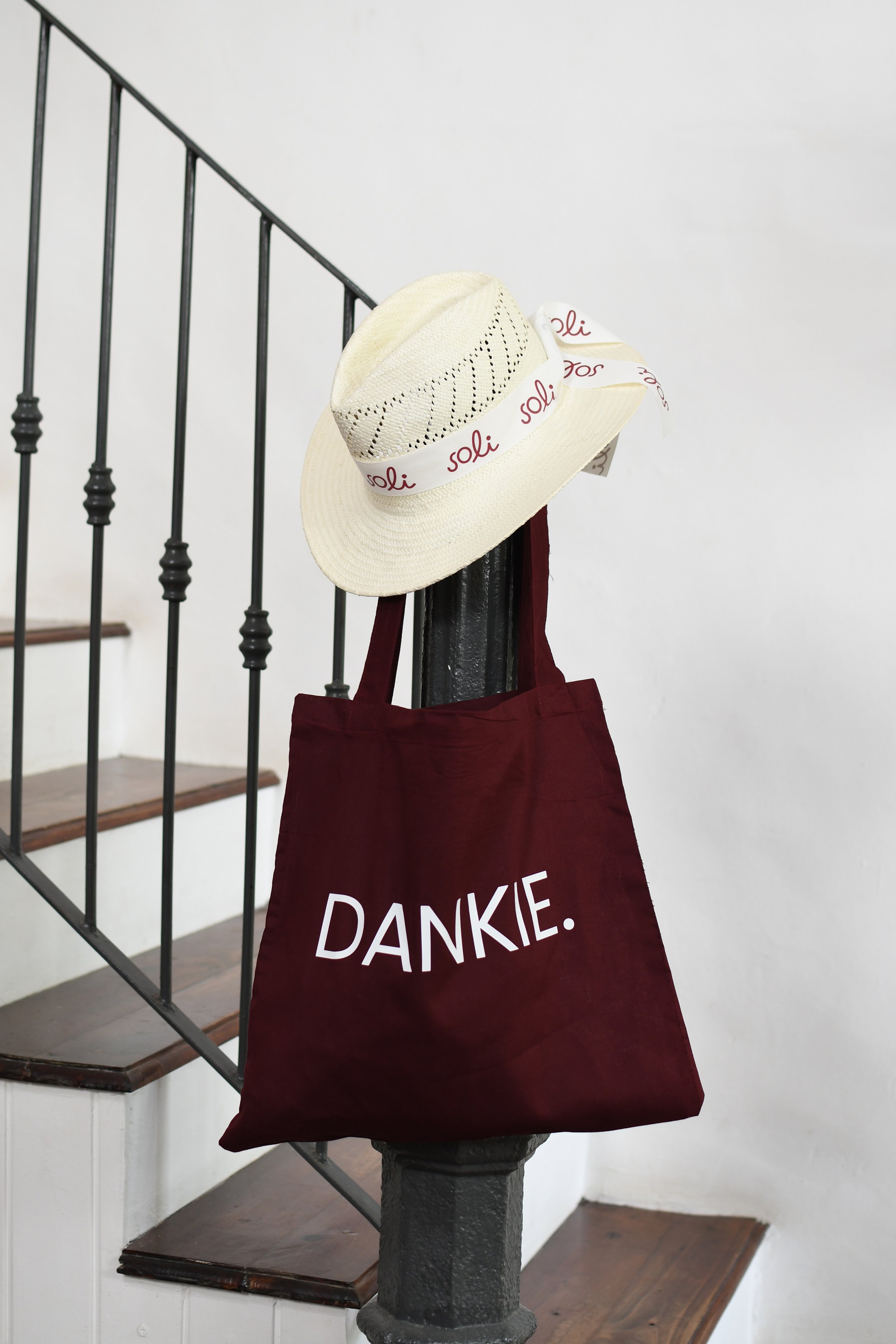 A beige straw hat with decorative holes and a "soli" branded ribbon, hanging on a stair railing next to a maroon tote bag with "Dankie." written on it.
