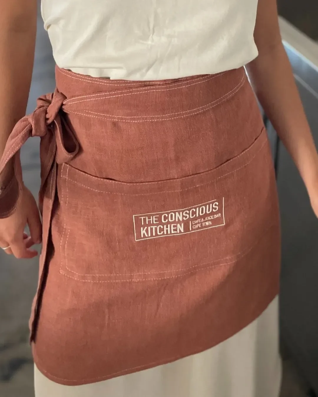 A close-up of a terracotta canvas half apron featuring a white printed logo for "The Conscious Kitchen," customized and handmade in South Africa.

