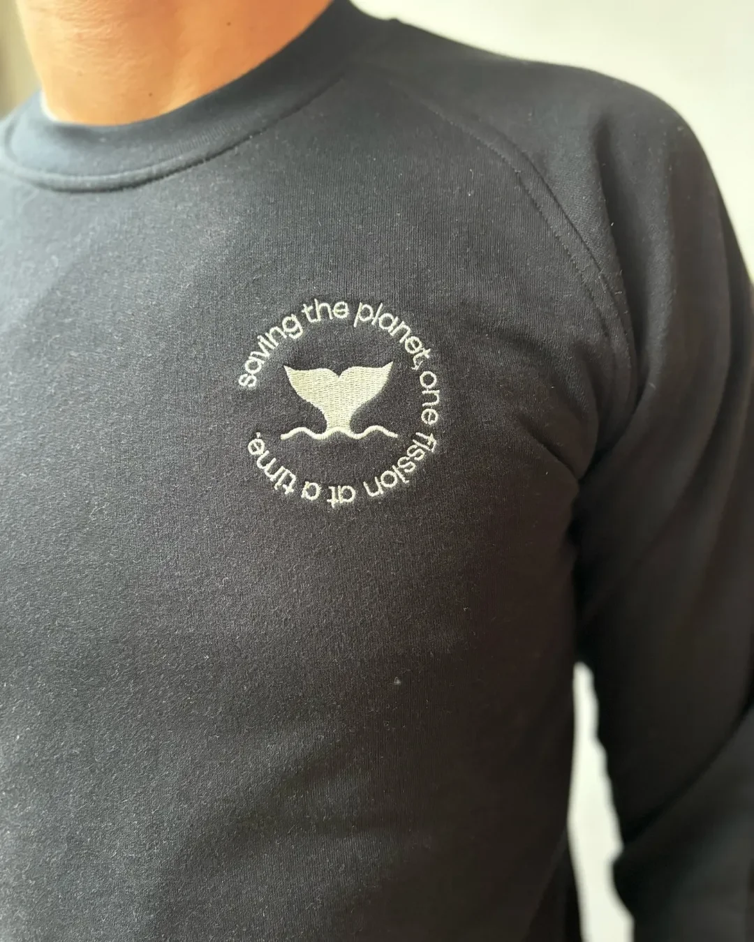 A close-up of a premium black 100% cotton sweater featuring a white embroidered circular whale tail logo, handmade by local artisans in Cape Town by Soli for Corporate gifts or merchandise.