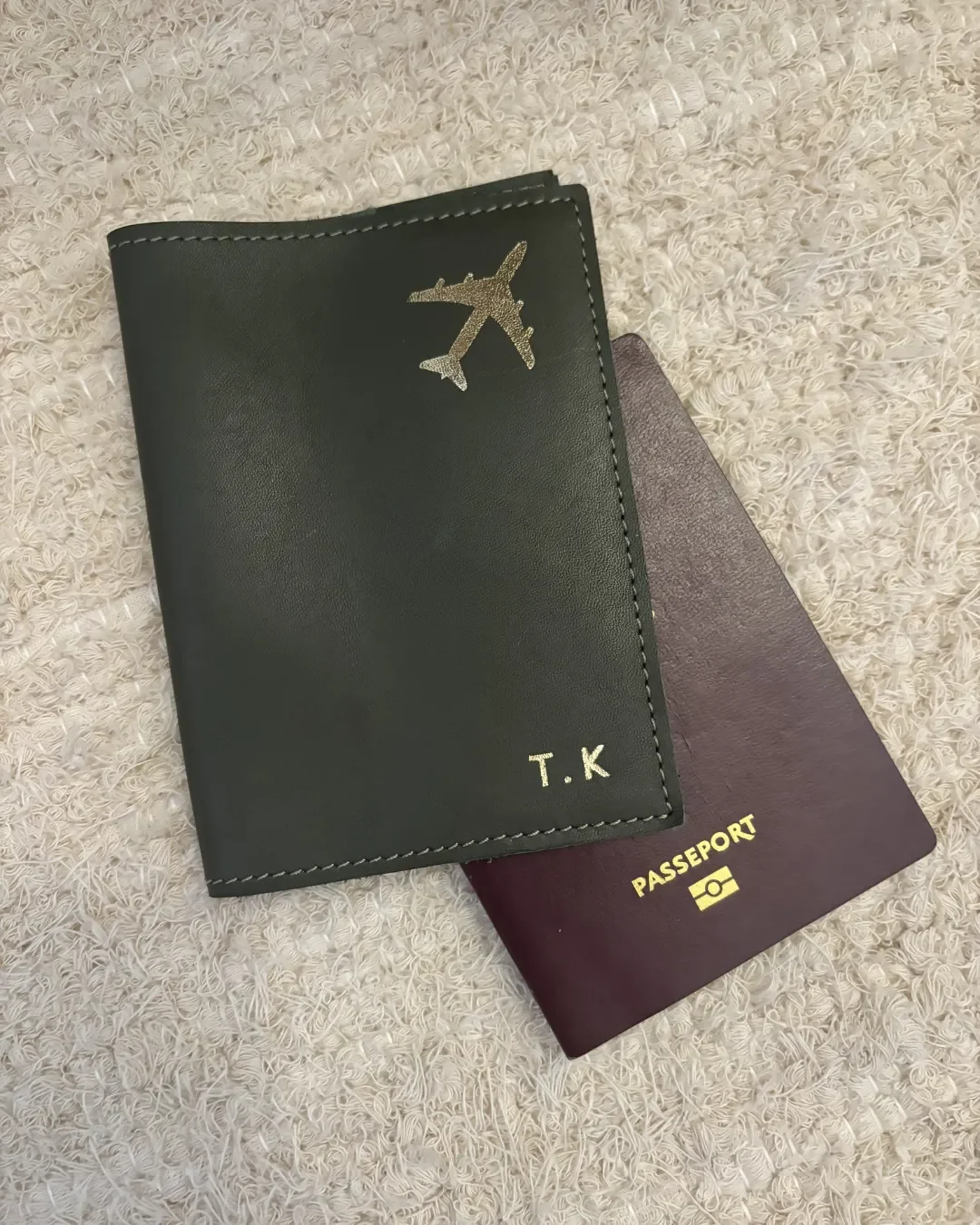 A custom olive green leather passport sleeve featuring a gold-foiled airplane icon and initials "T.K" resting on a white textured rug next to a French passport, Handmade by Soli in Cape Town for sustainable merchandise or corporate gifts.