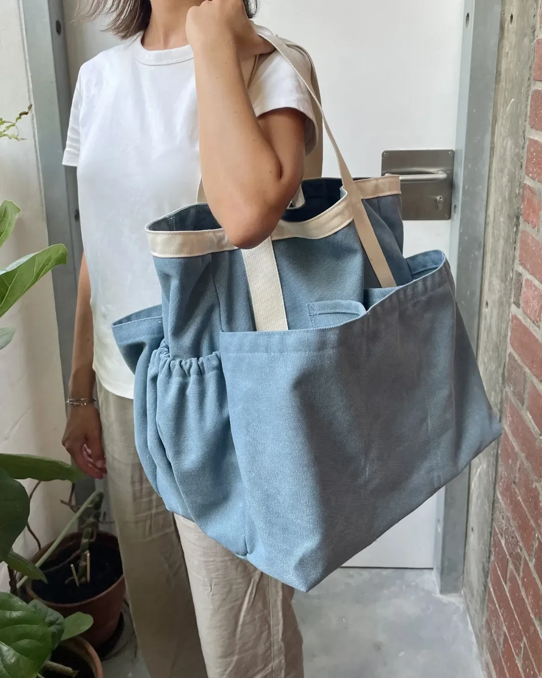 A person carrying a slate blue 100% canvas Market Bag featuring cream contrast handles and multiple spacious exterior pockets, Handmade by Soli in Cape Town for merchandise or corporate gifts.