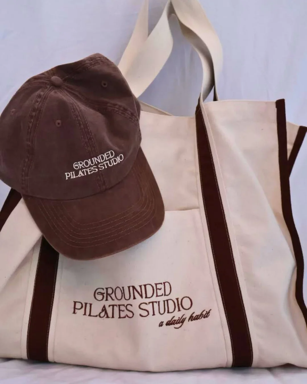 A cream-colored heavy canvas Diesel Bag featuring brown contrast handles and 'GROUNDED PILATES STUDIO' embroidery next to a matching brown cap, Handmade by Soli in Cape Town for merchandise or corporate gifts.
