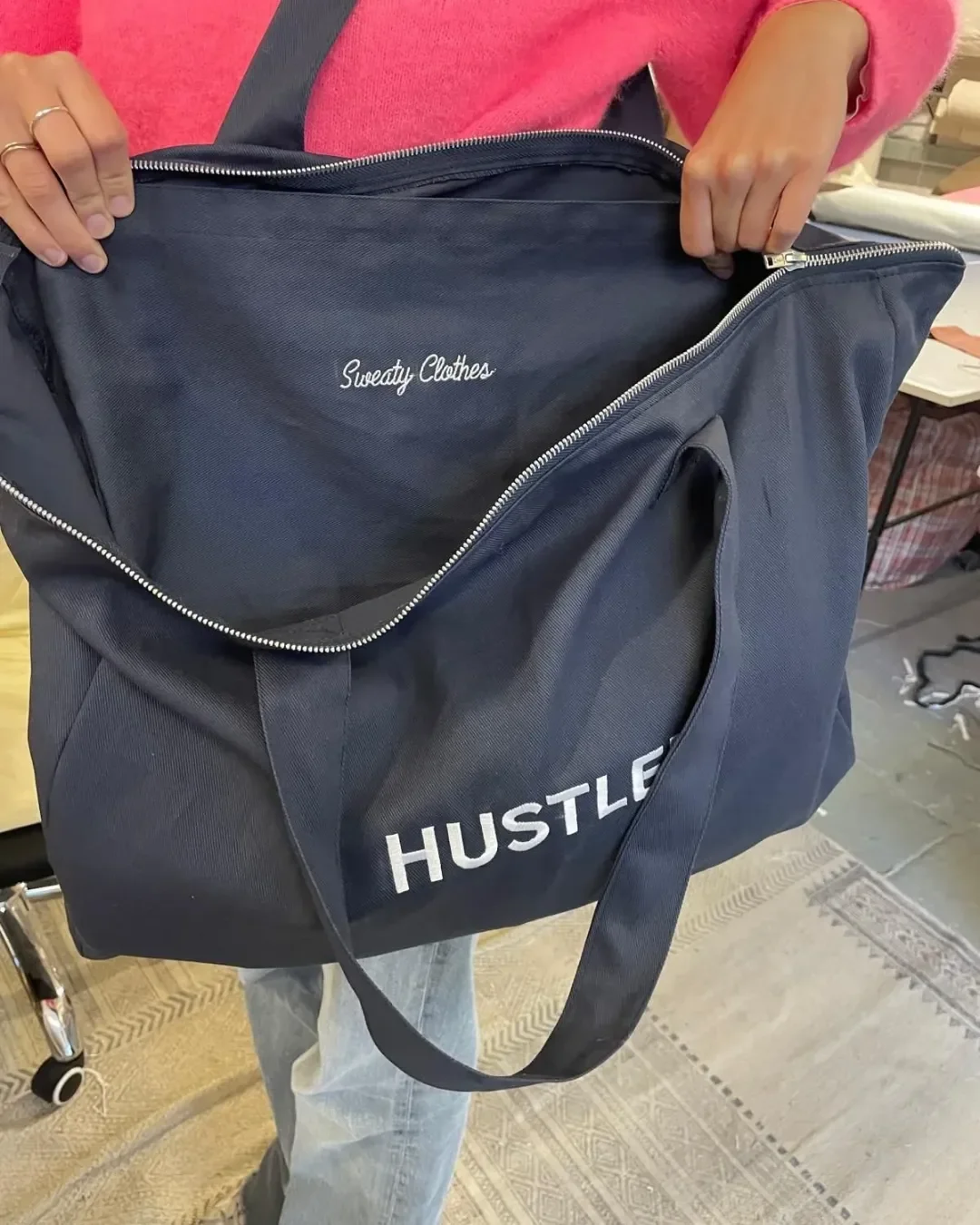 A person opens a large navy blue canvas zip tote bag branded with "HUSTLER", revealing an inner pocket printed with "Sweaty Clothes" in white cursive lettering for custom merchandise.