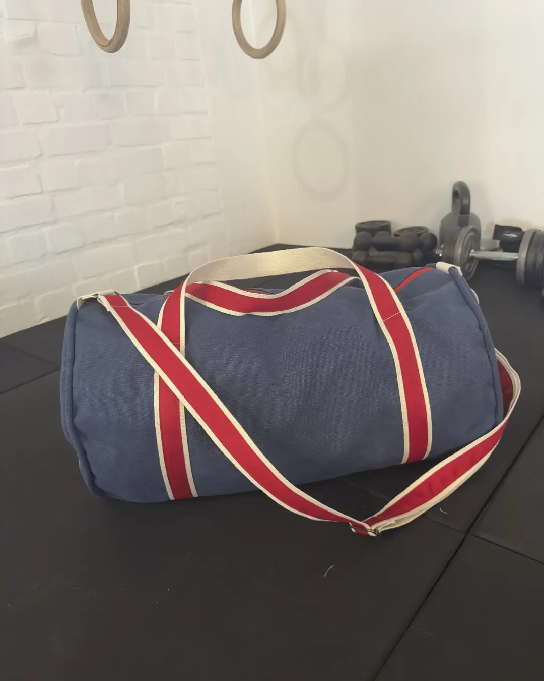 A blue canvas gym duffel bag with red and white striped straps resting on a black workout mat in a gym setting, handmade by Soli in Cape Town for sustainable retail merchandise.