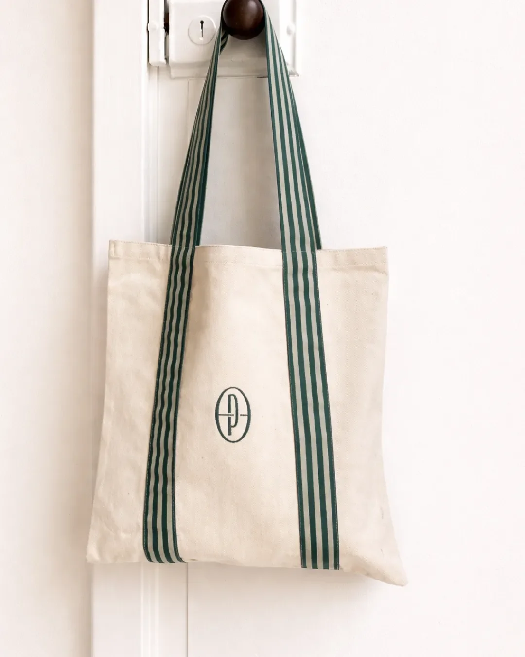 A cream-colored premium bull denim P-Bag hanging from a door handle, featuring a minimalist dark green oval logo and custom striped dark green and cream woven handles, handcrafted by local artisans in Cape Town for Soli.
