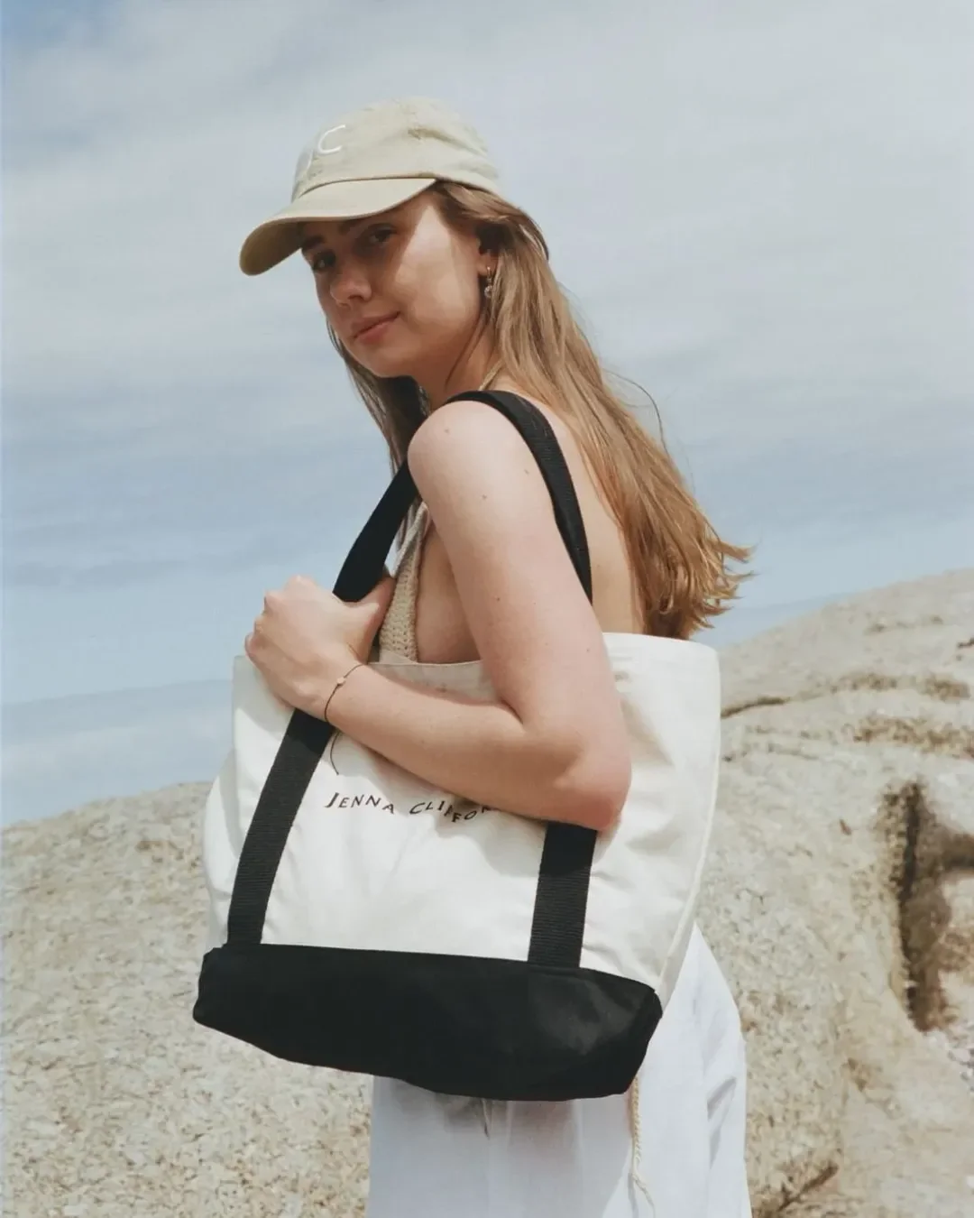 A woman wearing a beige cap and holding a custom Milano Tote bag in cream and black canvas with Jenna Clifford branding, handmade by Soli's artisans in Cape Town for merchandise or corporate gifts.