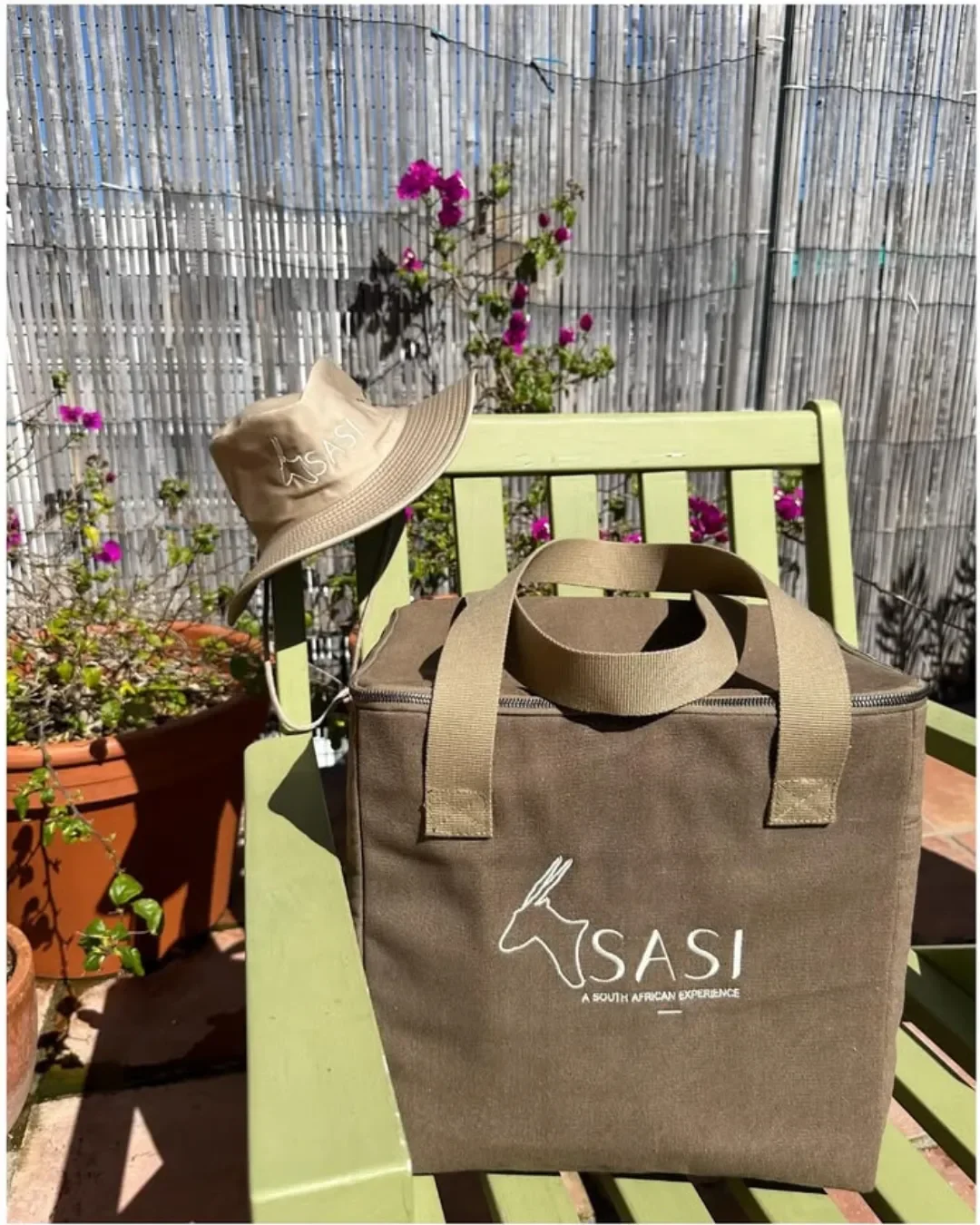 Brown heavy canvas cooler bag with custom white embroidered SASI logo and matching bucket hat on a green garden chair. Handmade by Soli in Cape Town for sustainable merchandise or corporate gifts.
