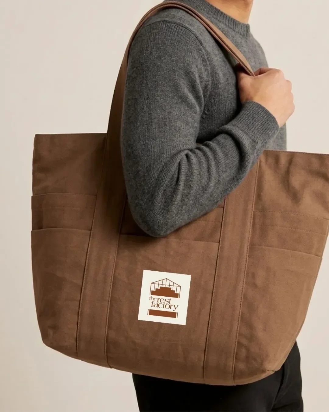 A large brown canvas tote bag featuring a white square printed logo for The Rest Factory, carried over the shoulder of a person wearing a grey knit sweater for custom brand merchandise.