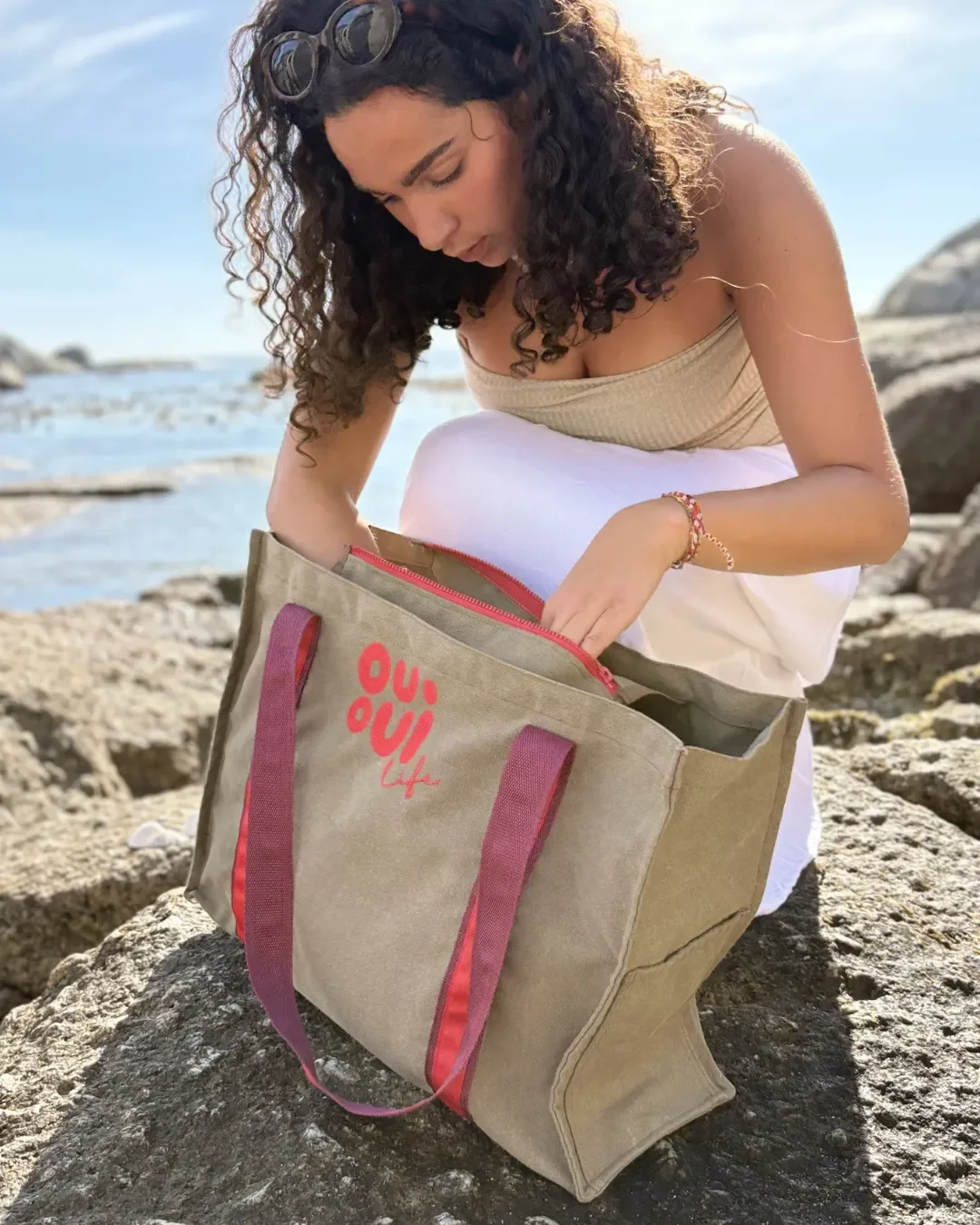 Person searching through a buckskin washed canvas Fire Bag with red vintage-style handles at the beach. Handmade by Soli in Cape Town for sustainable merchandise or corporate gifts.