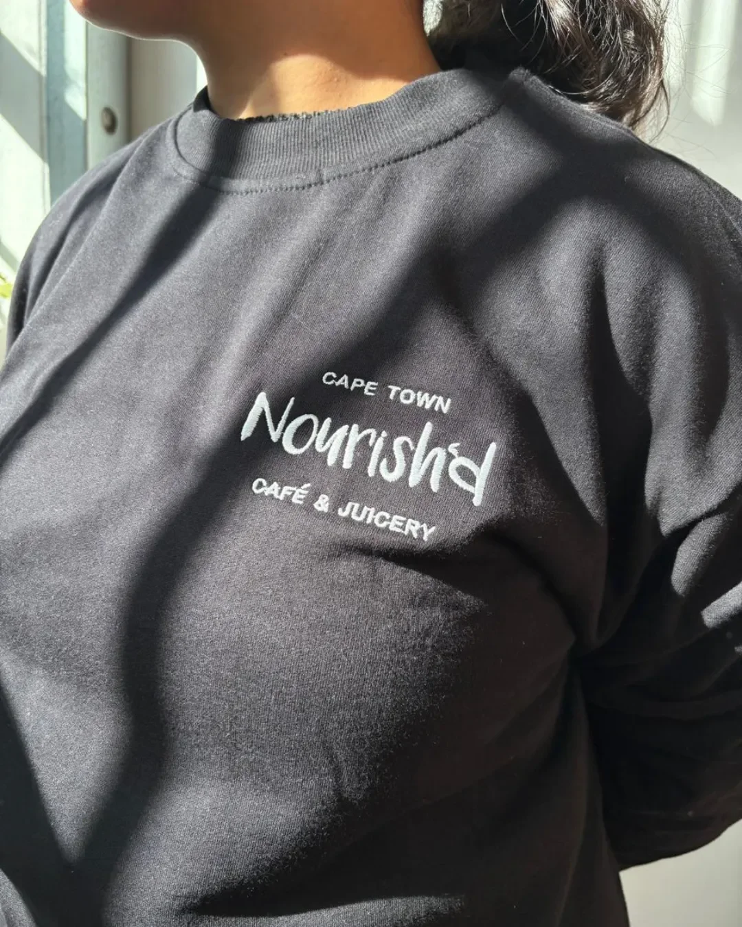 Close-up of a custom black crewneck sweatshirt featuring white chest embroidery that reads "CAPE TOWN Nourish'd CAFÉ & JUICERY", made by Soli in Cape Town.