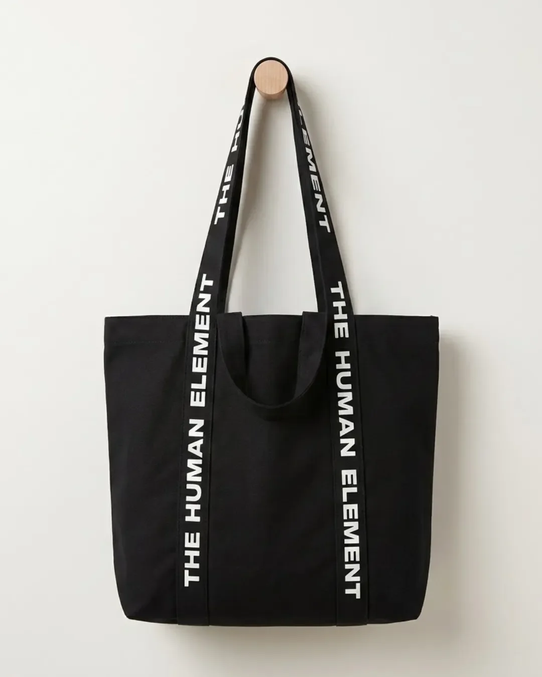 Black dual carry canvas tote bag hanging on a minimalist wooden wall peg showing long shoulder straps and short carry handles, Handmade by Soli in Cape Town for branded merchandise or corporate gifts