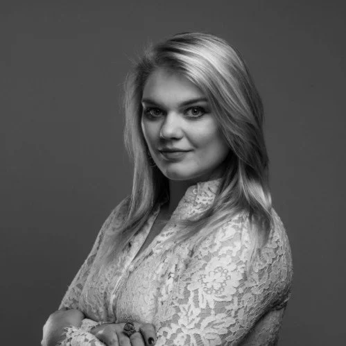 Black and white portrait of a woman with long hair and a lace top looking at the camera with a neutral expression.