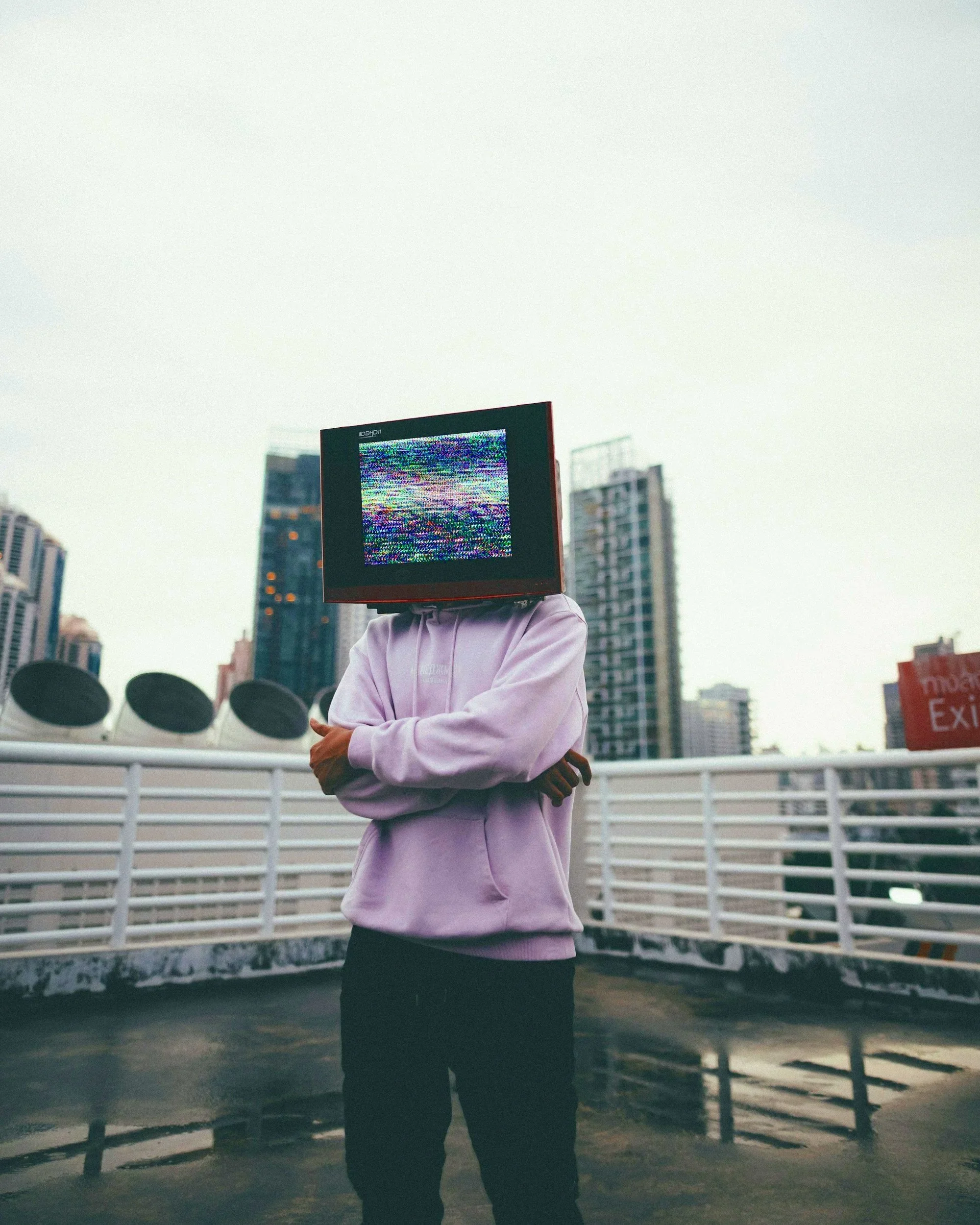 Person standing outdoors in front of city buildings, wearing a pink hoodie with a vintage television for a head showing static.