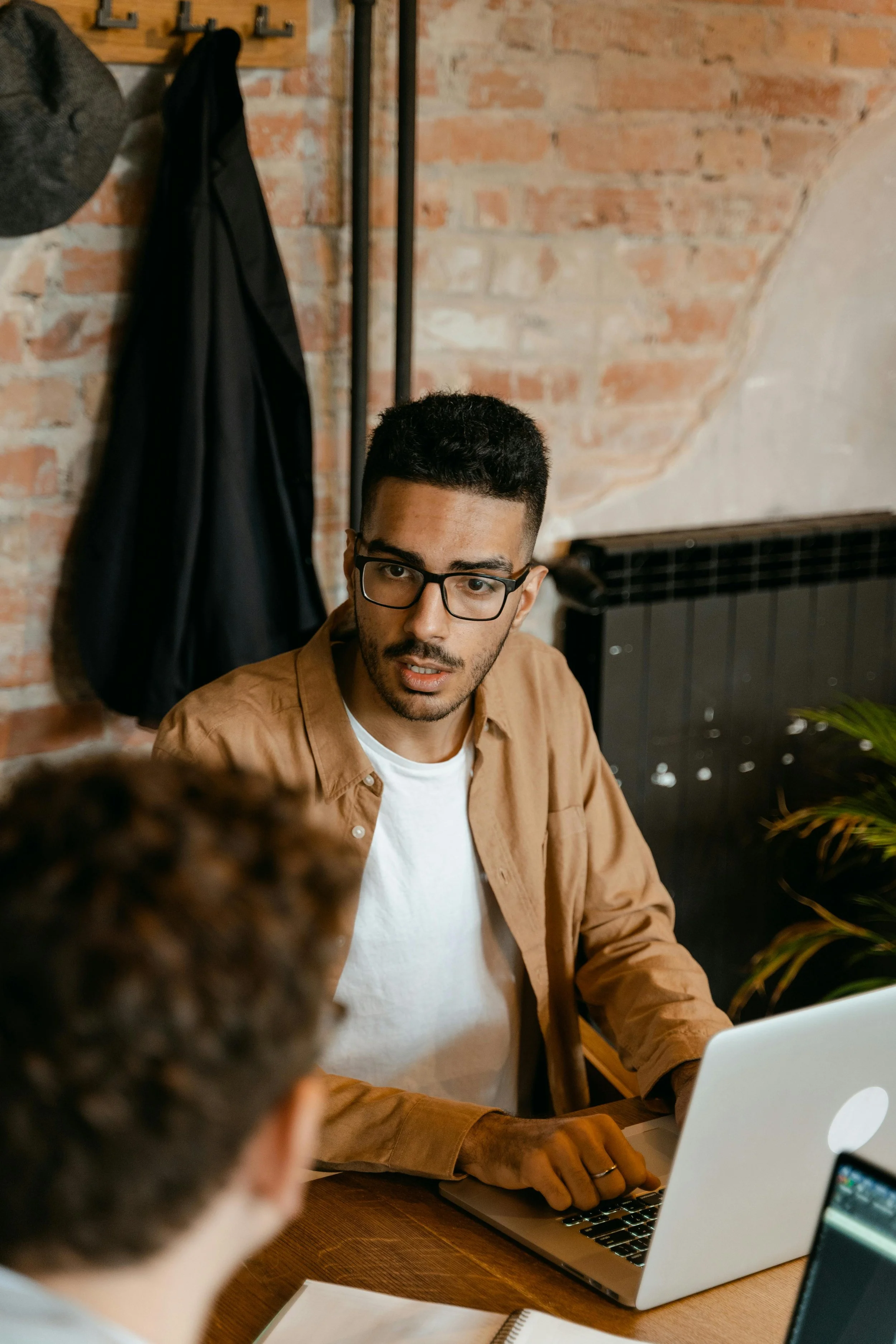 A man with black glasses, short dark hair, and a light brown shirt working on a laptop at a wooden table in a brick-walled room.