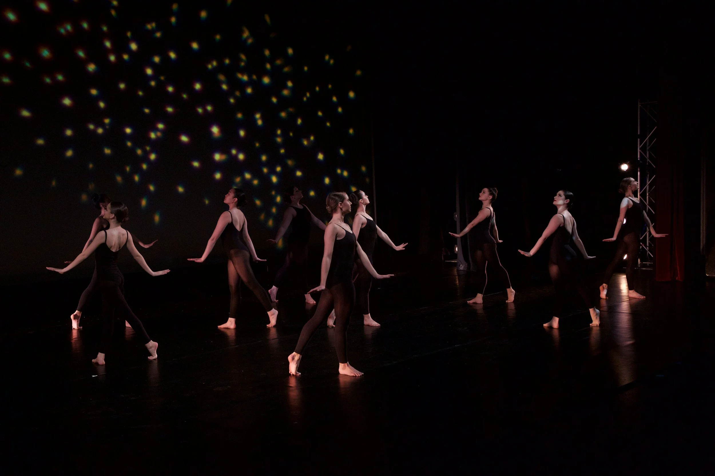 A group of dancers performing on stage in black costumes with a dark background. Light patterns are projected on the wall behind them.